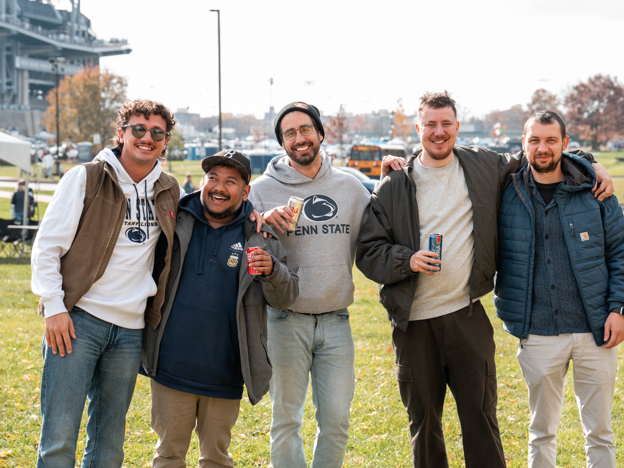 Five diverse young men standing outdoors on a grassy field, smiling and holding drinks, with a Penn State sweatshirt and casual clothing, in a park-like setting during fall.