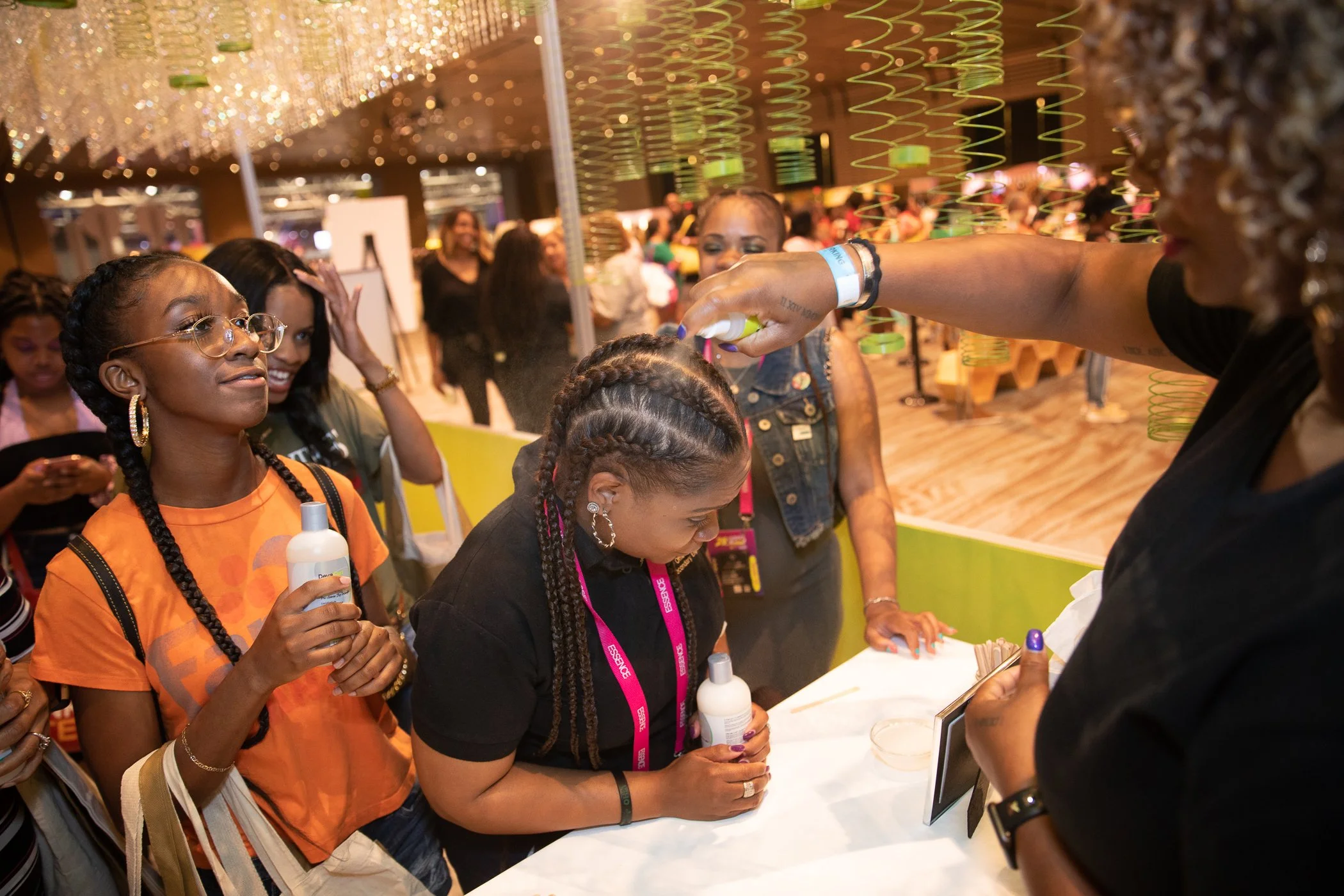 A group of women at a booth, with one woman applying lotion or cream to another woman's head while others watch, in a brightly lit indoor setting.