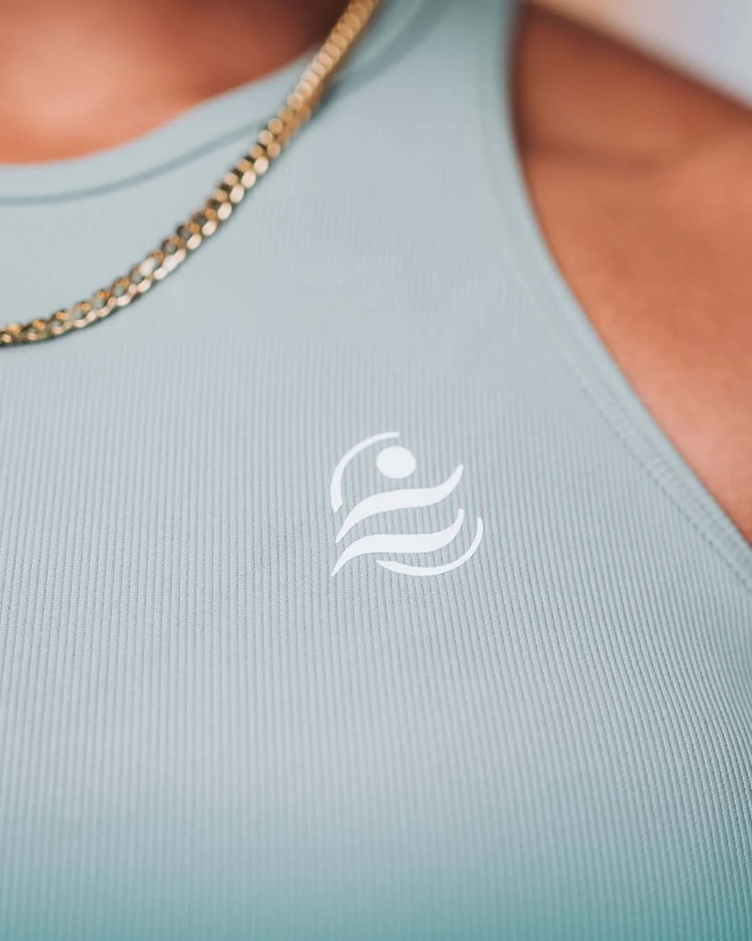 Close-up of a light blue athletic top with a white logo of a figure swimming, featuring a stylized wave and a circle representing the sun or moon, on the chest area. A gold chain necklace is partially visible around the neck.