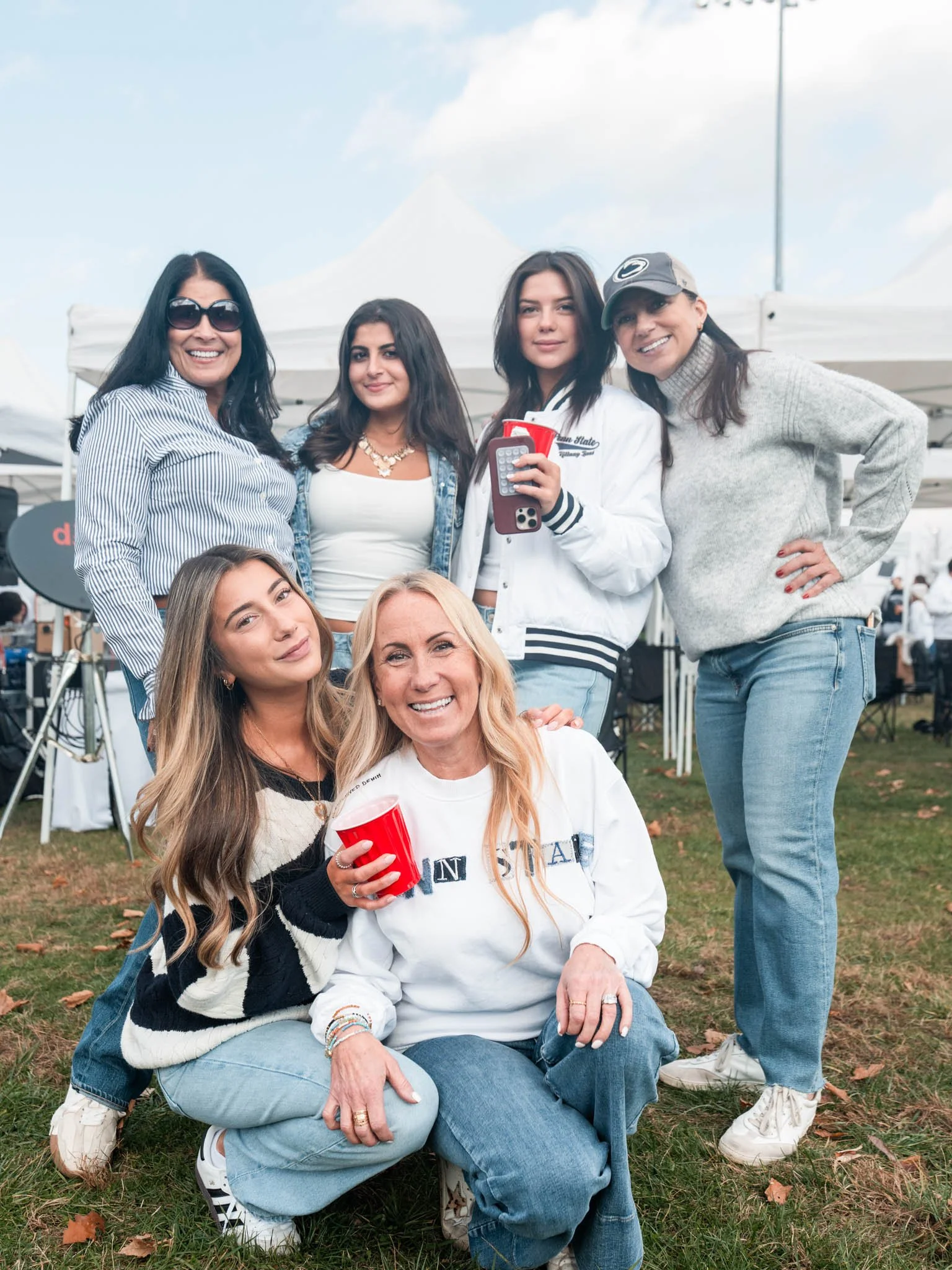 Group of six women smiling at an outdoor event, some holding cups and a phone, in front of white tents on grassy ground.