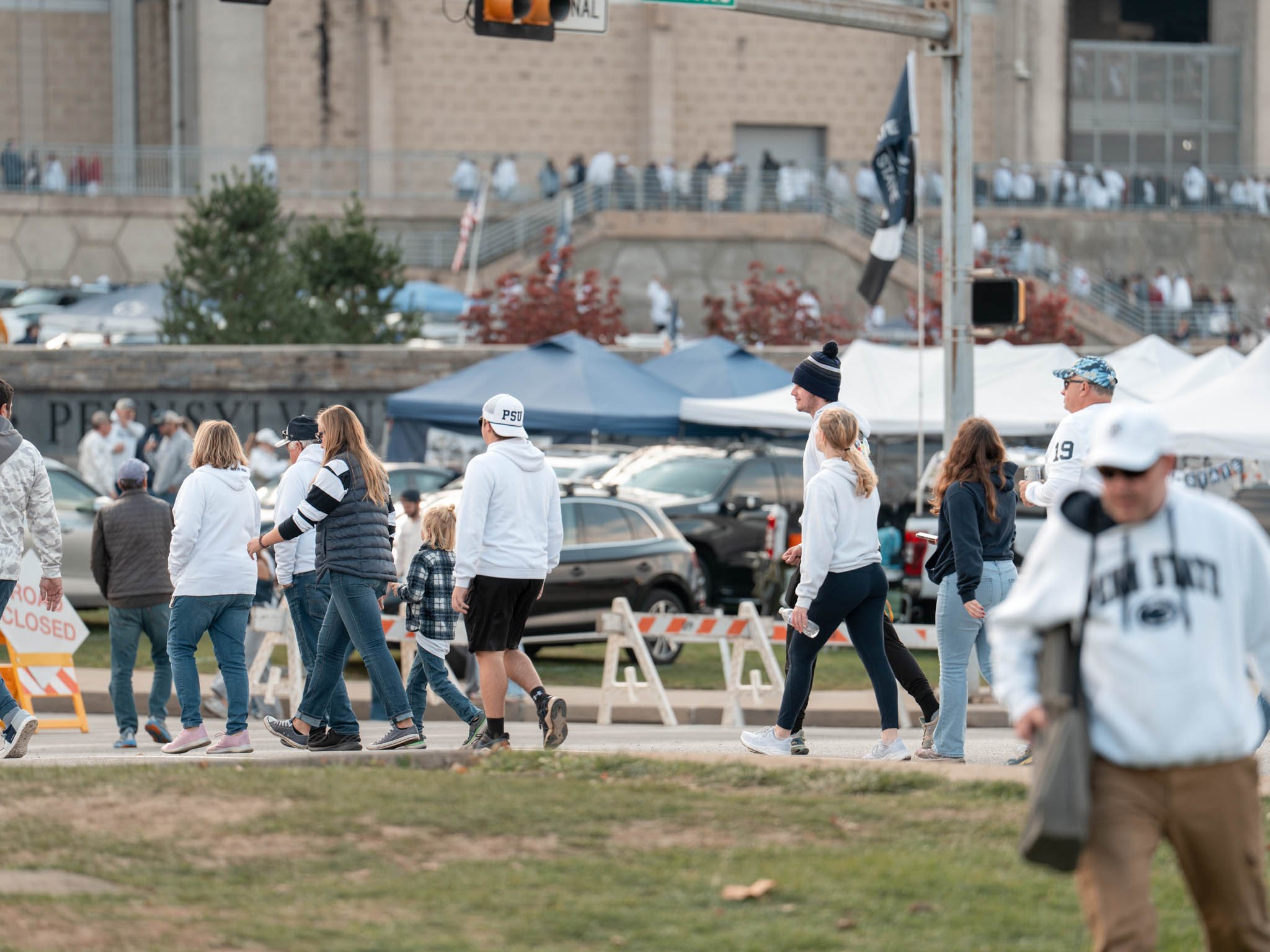 People walking outside near tents and parked cars during daytime, with a brick building and trees in the background.