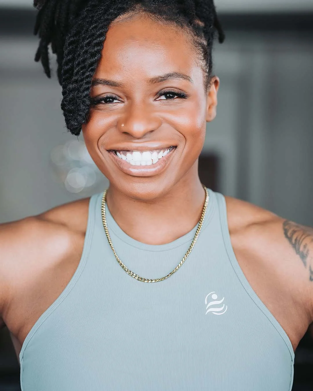 A smiling woman with short dreadlocks, wearing a light blue athletic top with a small logo, a gold chain necklace, and a nose piercing. She has a radiant smile, and there is a tattoo visible on her upper arm.