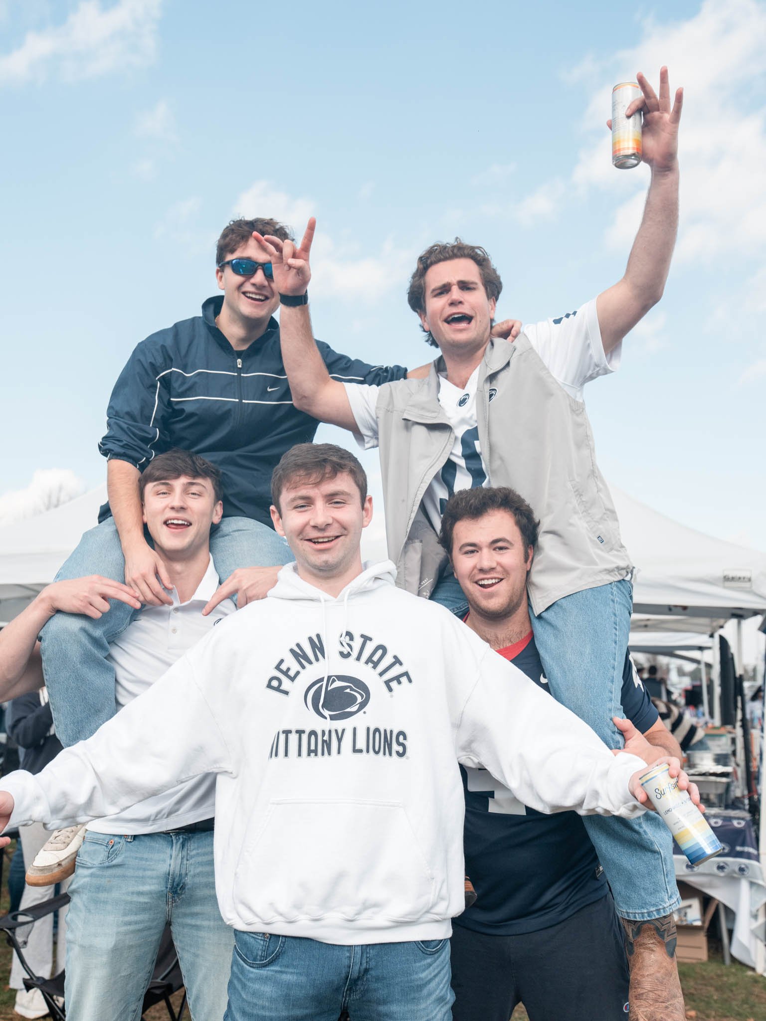 Group of five young men smiling and posing at an outdoor event under tents, with some holding cans, wearing casual clothing including a Penn State hoodie.