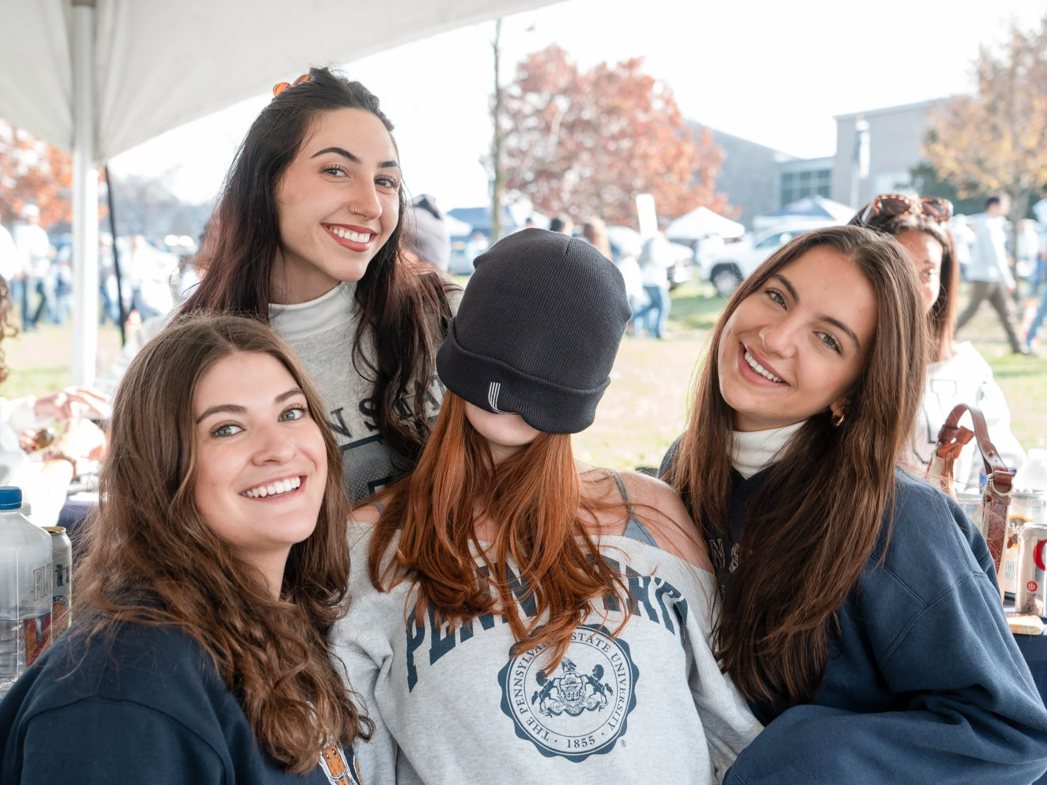 Group of five smiling young women at an outdoor event under a tent, with trees and other people in the background.