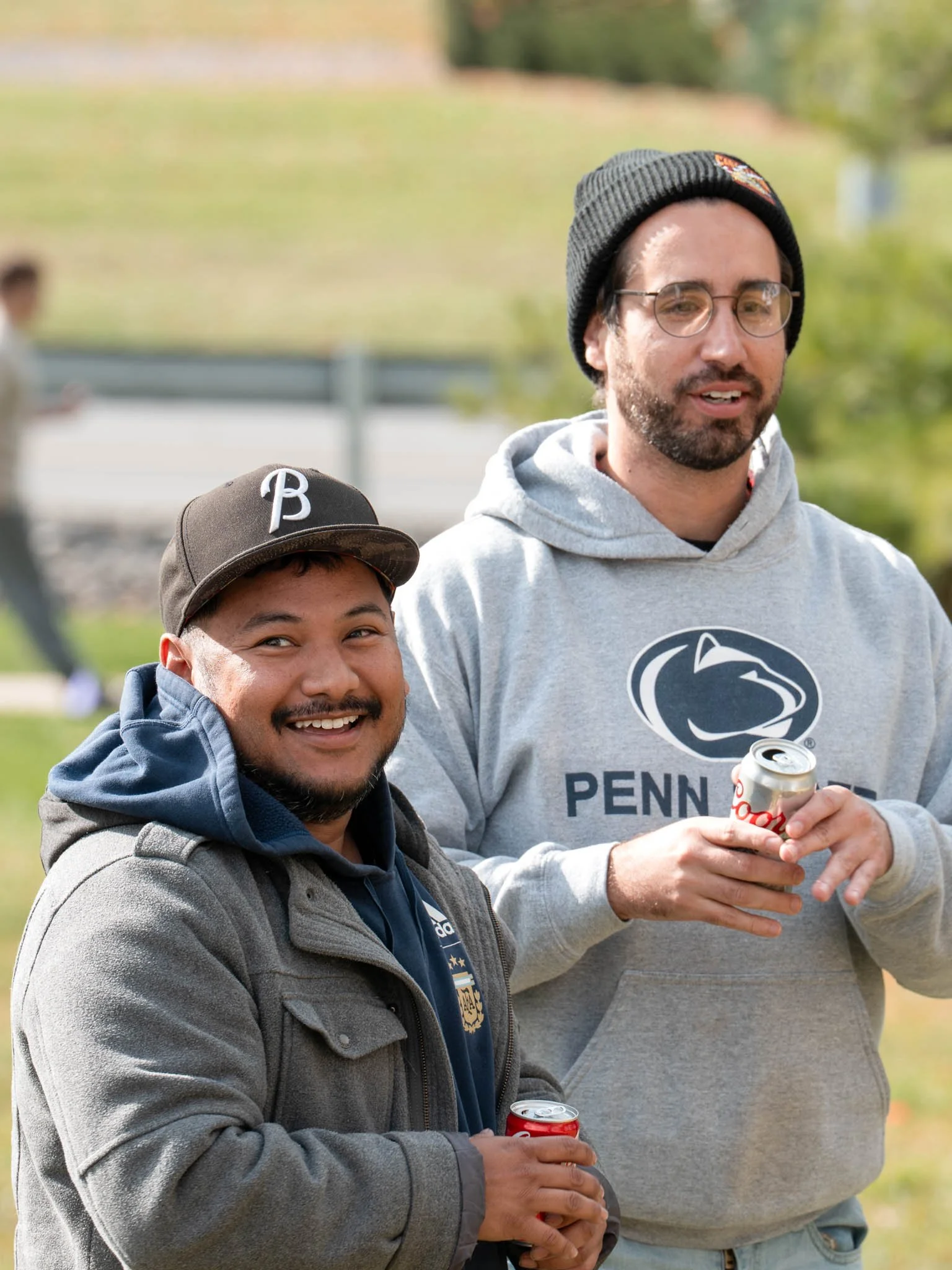 Two men standing outdoors, smiling and holding cans of soda, with a blurry person in the background. One wears a Penn State hoodie and a beanie, the other wears a cap and a jacket.
