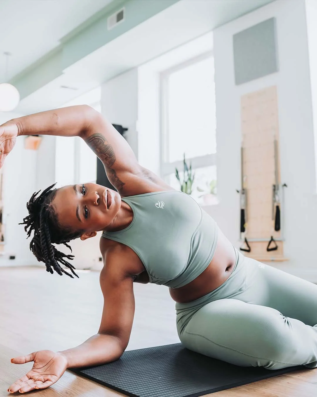 A woman with dreadlocks wearing a matching olive green sports bra and leggings performing a side-lying stretch or yoga pose on a gray exercise mat in a bright, modern home gym with large windows and workout equipment in the background.