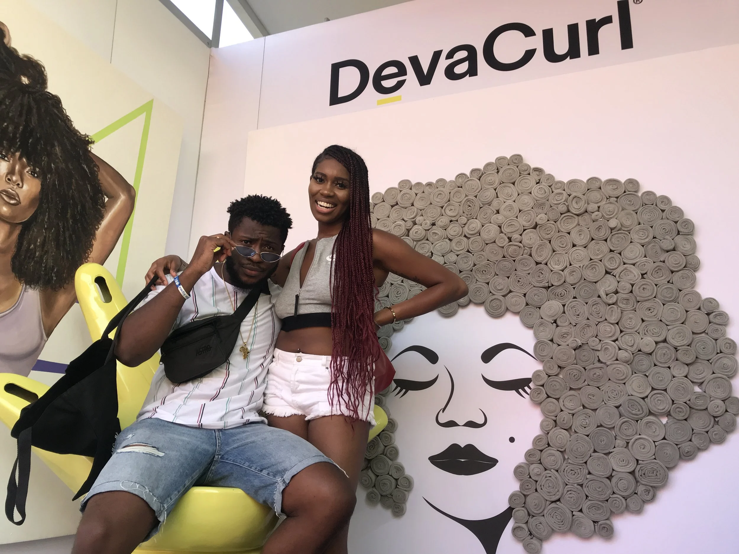 Two people posing in front of a wall art of a woman's face with large curly hair made of rolled paper. The person on the left is a man wearing a striped shirt, ripped denim shorts, sunglasses, and carrying a black bag. The person on the right is a wo