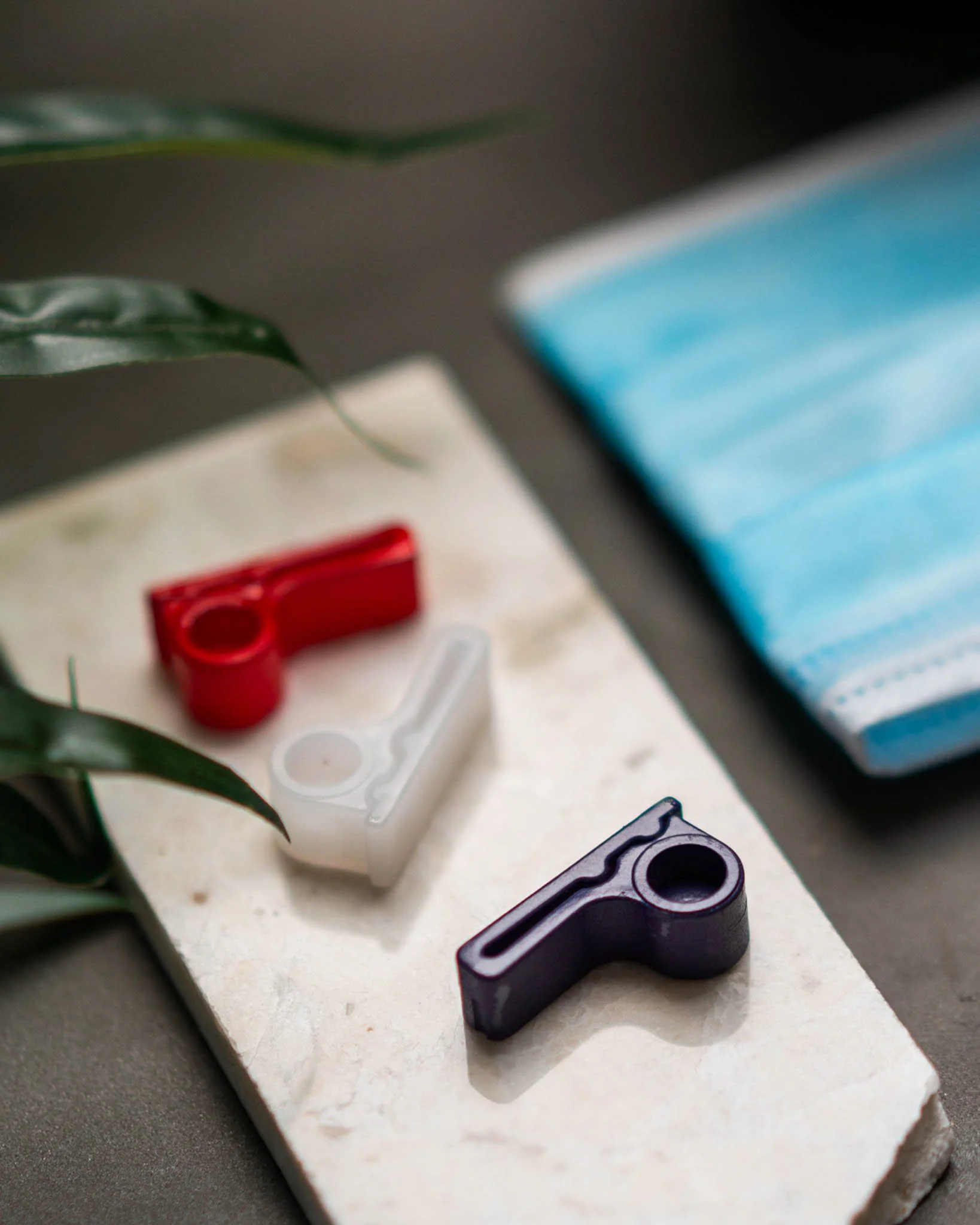 Three small, colored, camera-shaped objects in red, white, and black are arranged on a beige stone tile, with a plant and a blue face mask in the background.