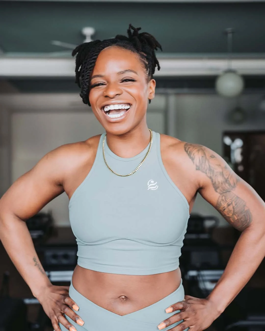 A woman with dreadlocks and tattoos on her arms, wearing a light gray athletic top and matching leggings, smiling with her hands on her hips in a gym.
