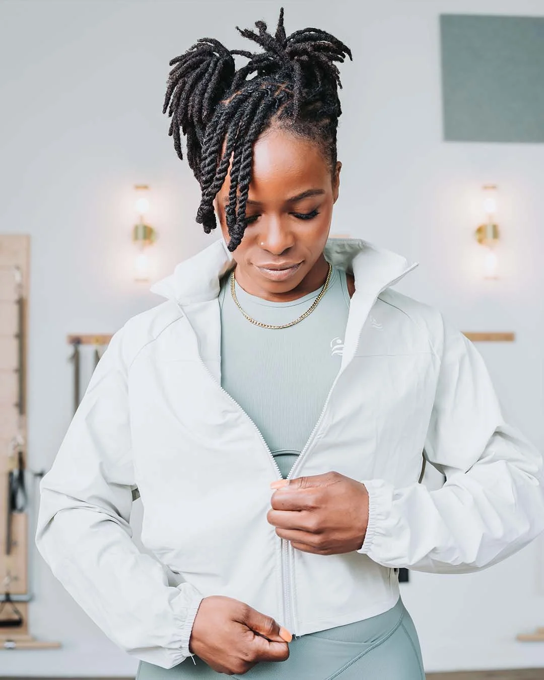 A woman with styled dreadlocks wearing a light-colored athletic outfit and a white zip-up jacket, standing in a bright room with modern lighting.