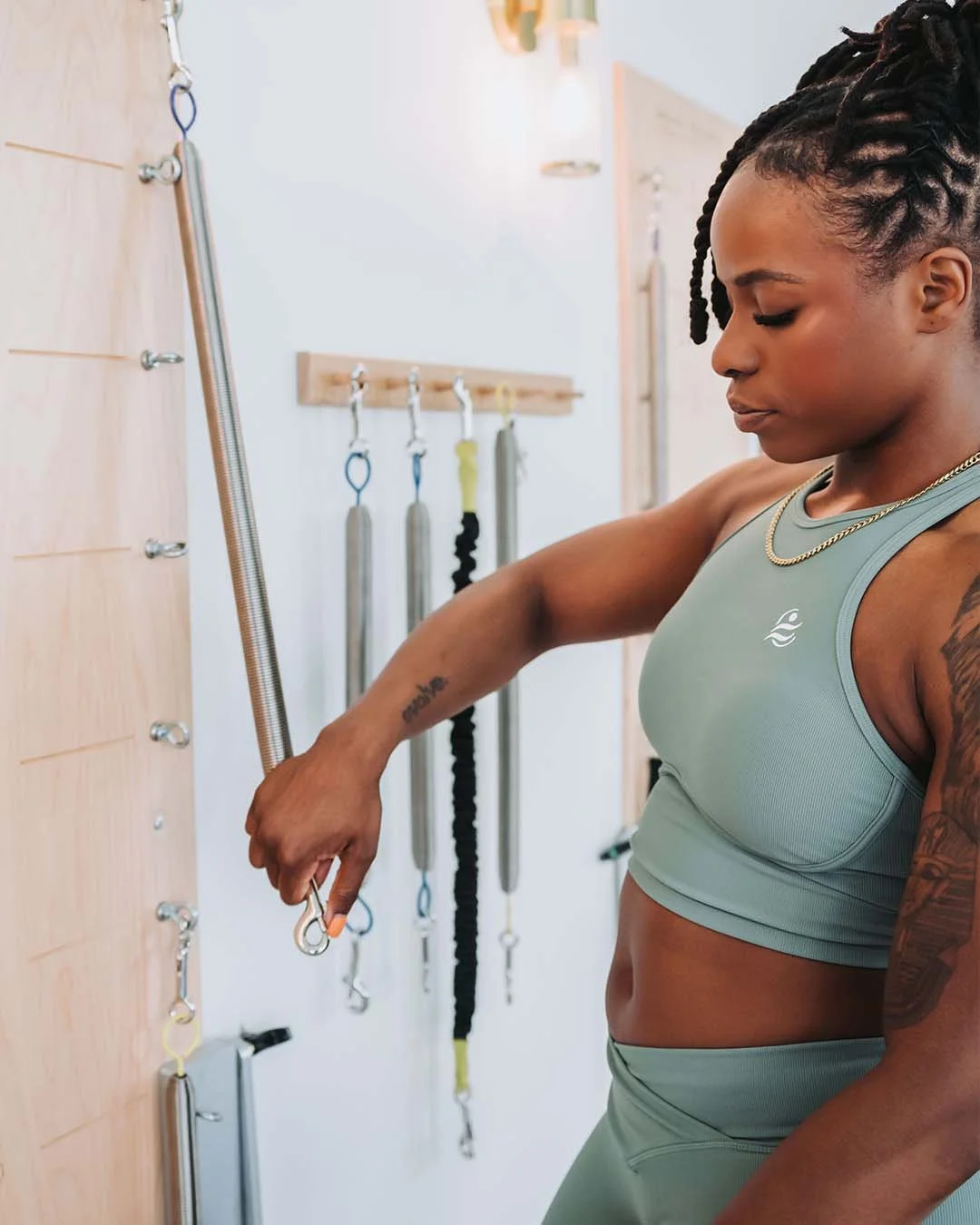 A woman with braided hair in athletic wear is grabbing a ring attached to a wall in a fitness studio, with various gym tools hanging on hooks behind her.