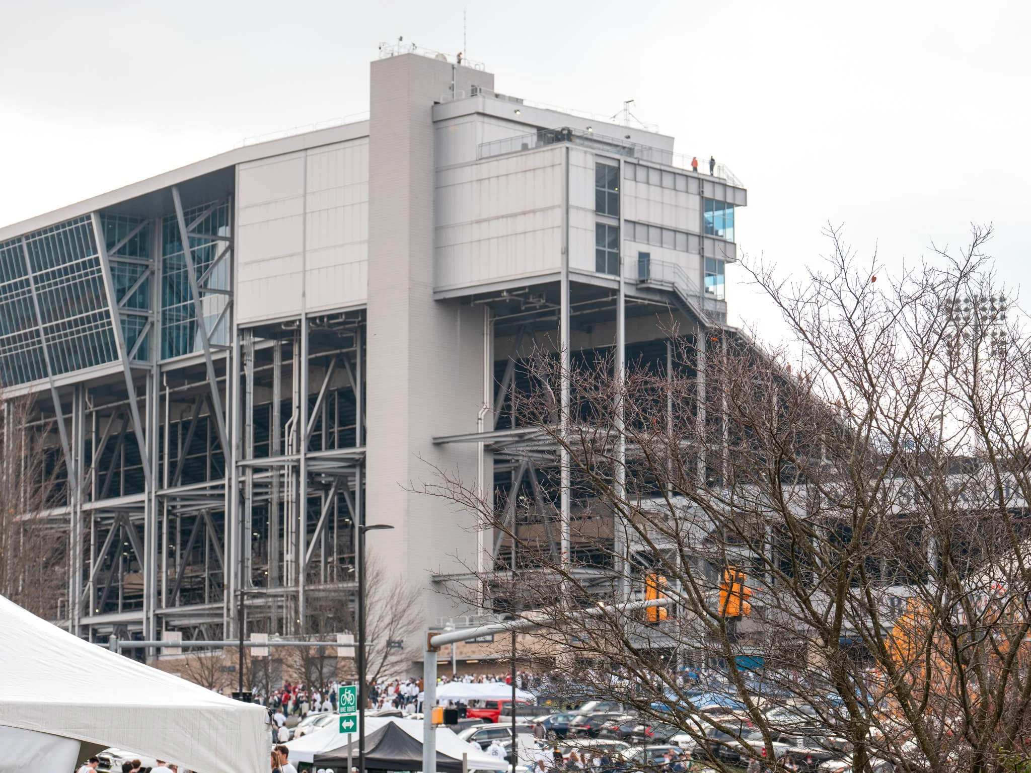 Modern multi-story parking garage with a metal framework, trees with bare branches in front, and tents at the base, with overcast sky.