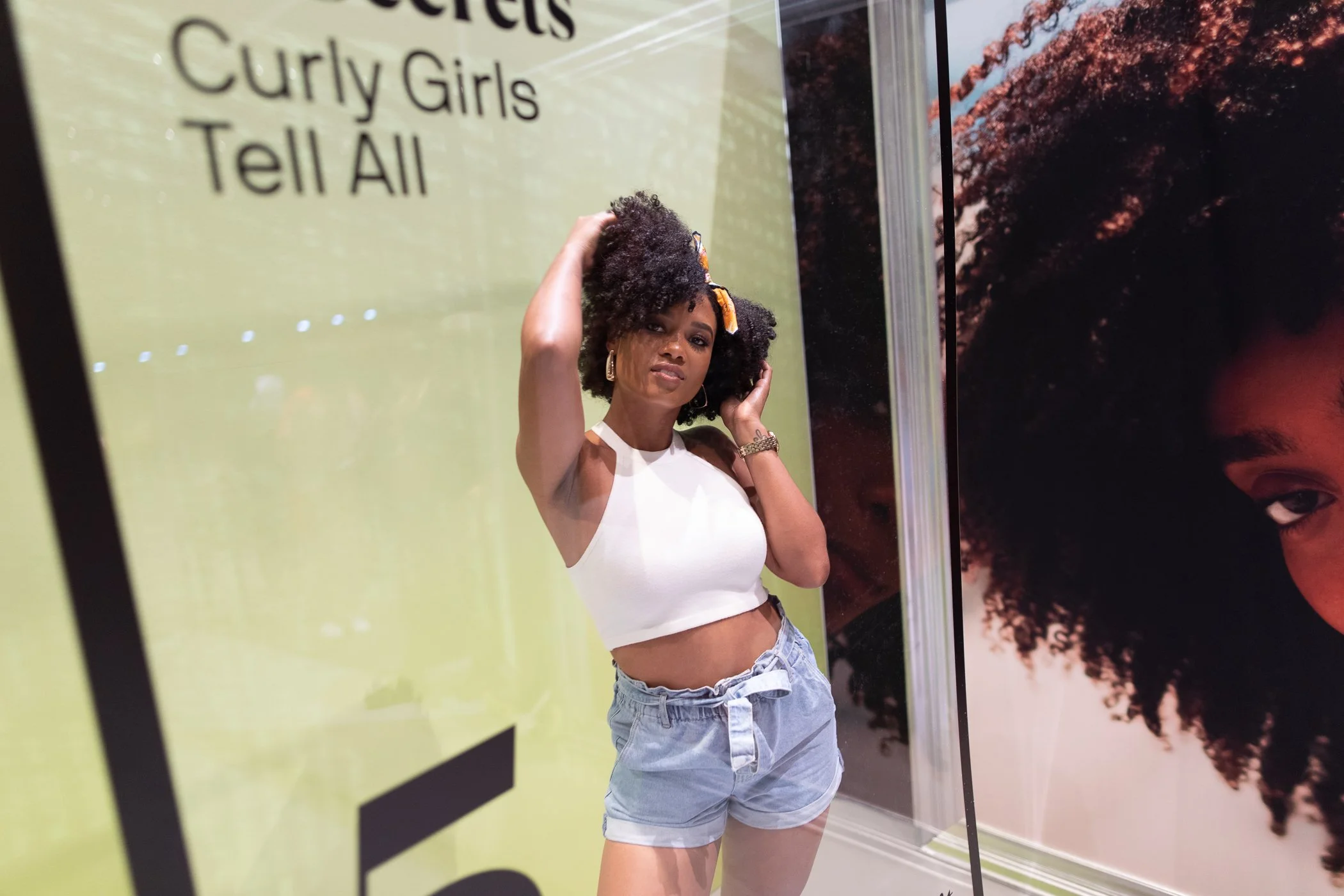 A woman with curly hair wearing a white sleeveless top and denim shorts posing in front of a yellow and black sign that says 'Curly Girls Tell All' and a large portrait of a woman with natural hair.