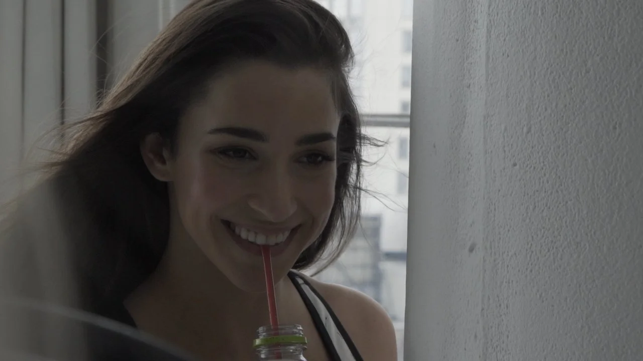 A young woman with long dark hair smiling while drinking from a bottle with a red straw by a window.