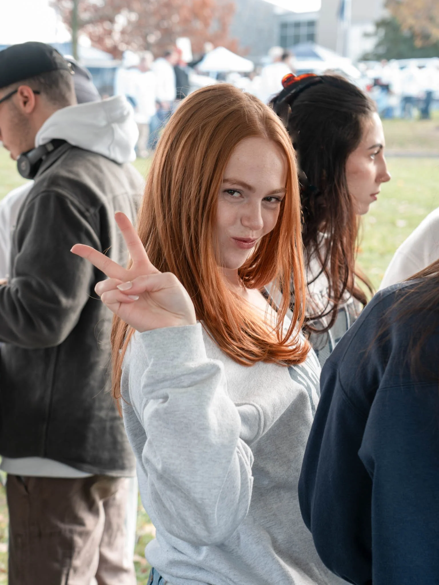 A red-haired woman making a peace sign and smirking at a camera, standing next to a dark-haired woman in profile, at an outdoor event with tents and other people in the background.