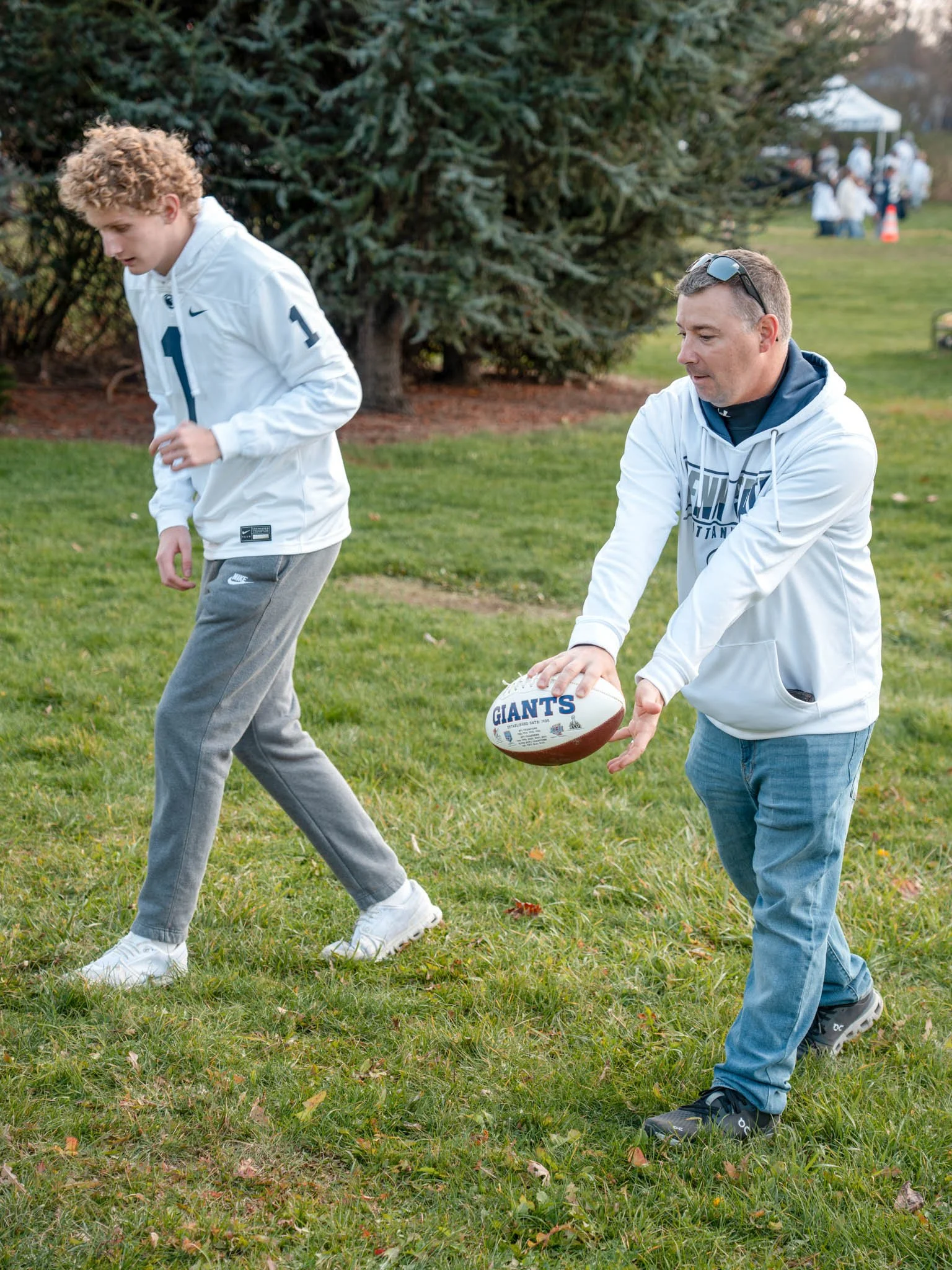 A man and a teenage boy practicing football outdoors on a grassy field with trees in the background. The man is holding a football with the word 'Giants' on it, preparing to throw, while the boy looks down, wearing a white hoodie and gray sweatpants.