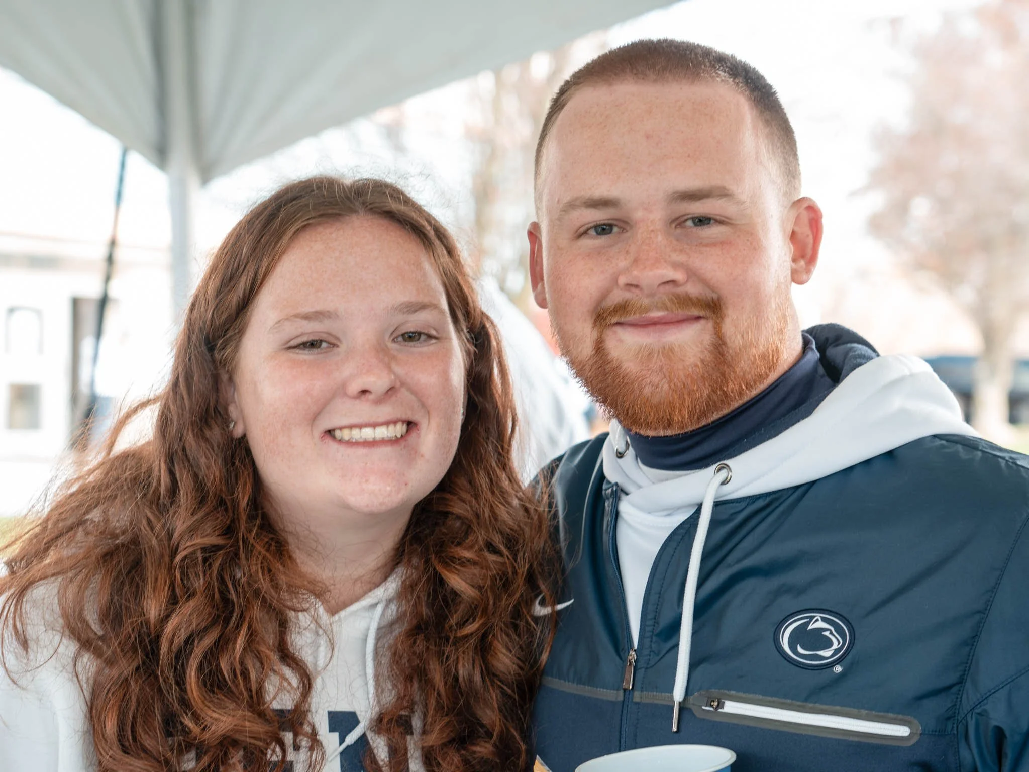 A young woman with long red hair and a young man with short red hair and beard smiling outdoors, standing close together under an umbrella.