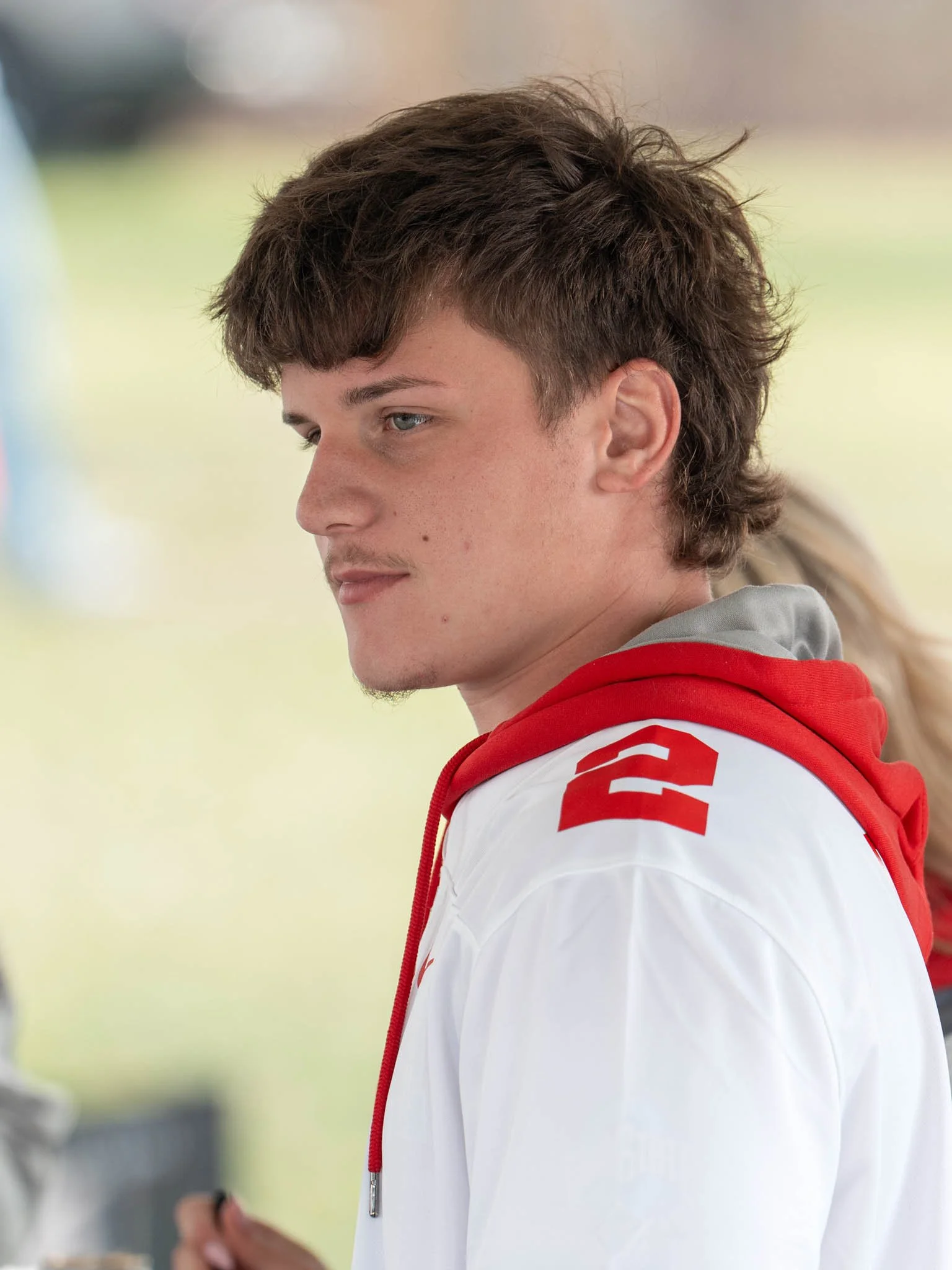 A young man with brown, curly hair wearing a white and red jacket, looking to the side.