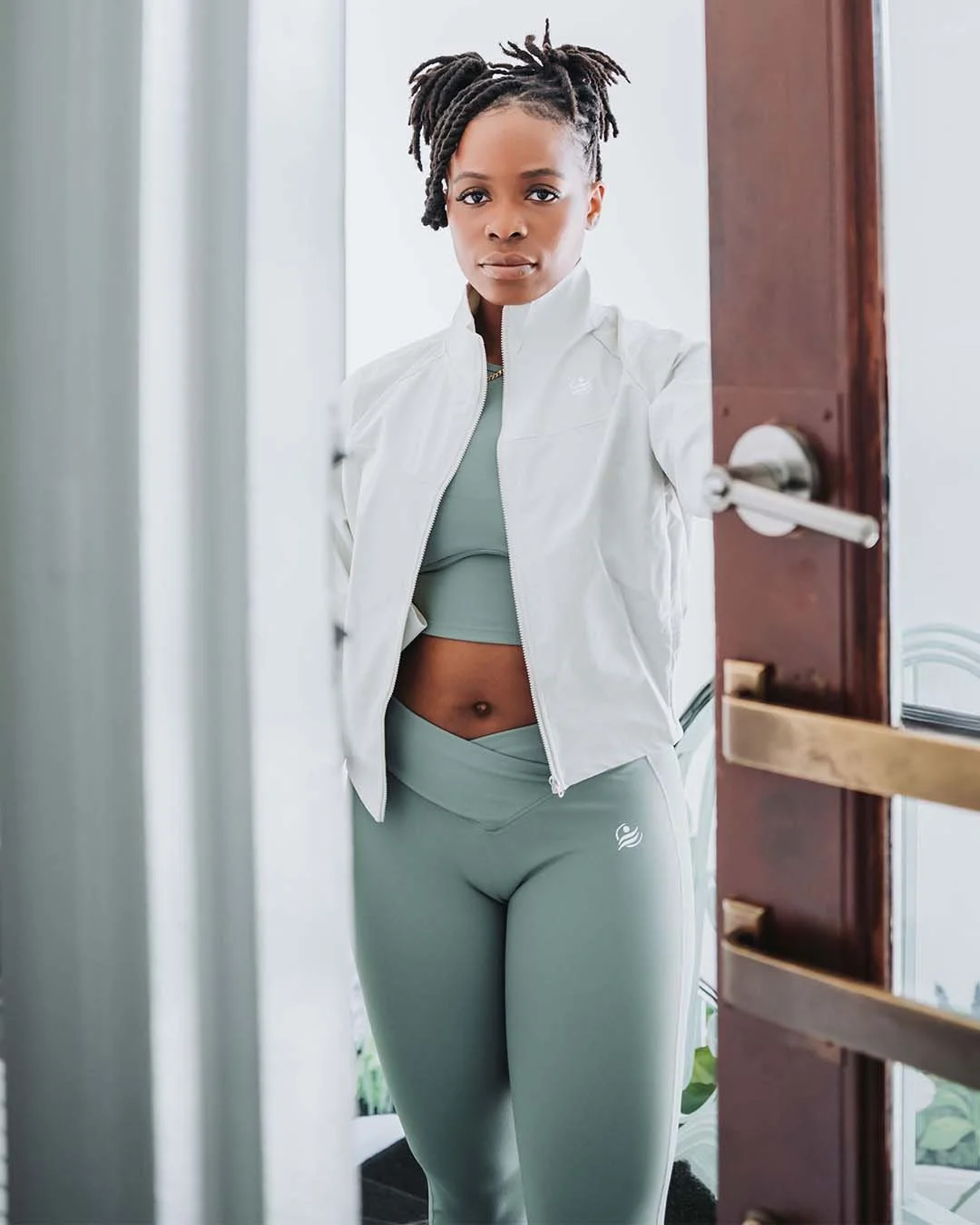 A woman with dreadlocks is taking a mirror selfie in a workout outfit, wearing a light gray zip-up jacket and matching leggings.