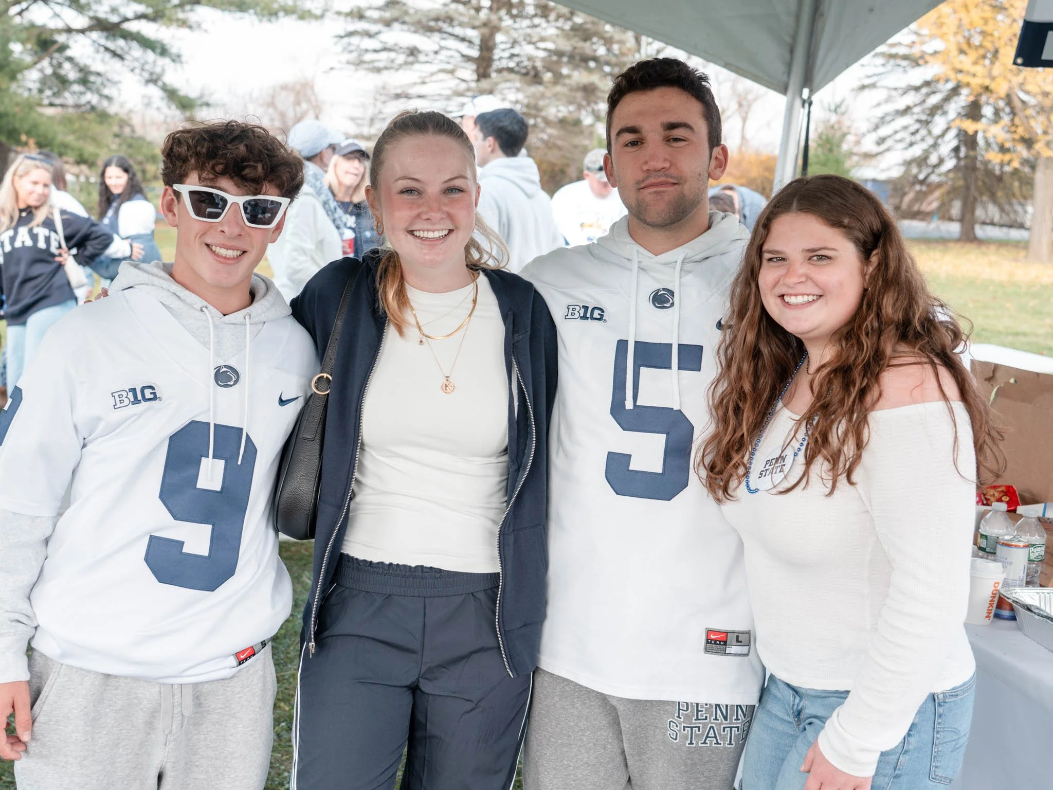 Four young people standing together outdoors, smiling, with Penn State apparel and casual clothing, at an event with tents and other people in the background.