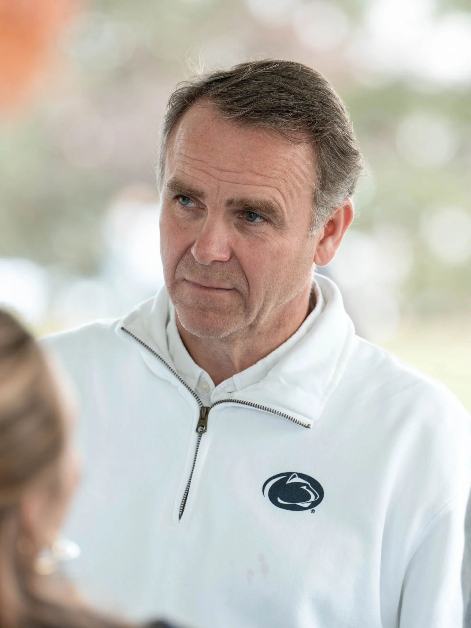 A middle-aged man with gray hair and blue eyes wearing a white Penn State quarter-zip pullover, appears to be engaging in conversation with a woman.