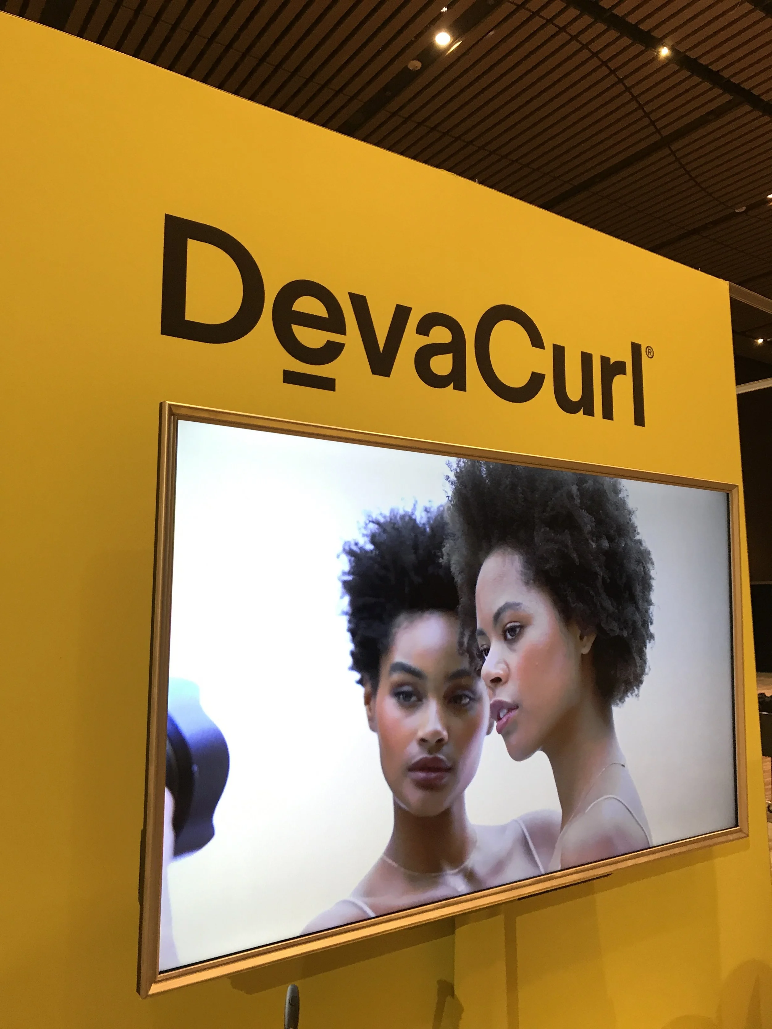 A digital screen displaying two women with natural hair, posing closely for a photo at a DevaCurl event, with yellow branding background.