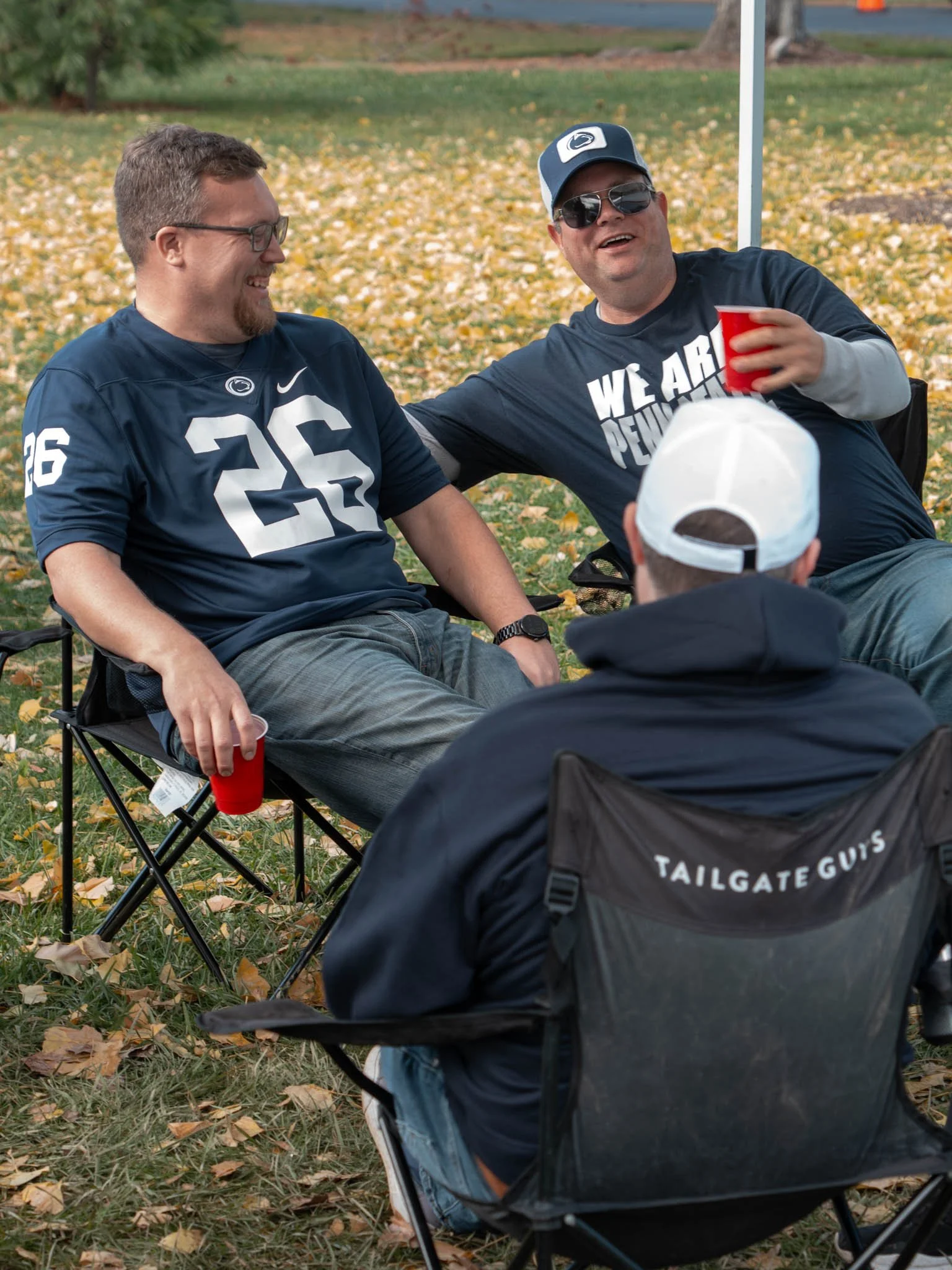 Three men sitting outdoors on folding chairs on fallen leaves, enjoying drinks and conversation, with fall foliage in the background.