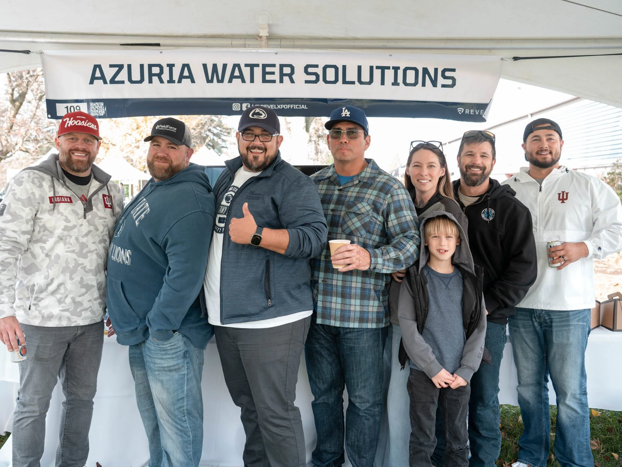 Group of seven people, including six men and one boy, standing under a white tent with a banner reading 'AZURIA WATER SOLUTIONS'. They are dressed casually, some wearing hats or hoodies, and are smiling at the camera.