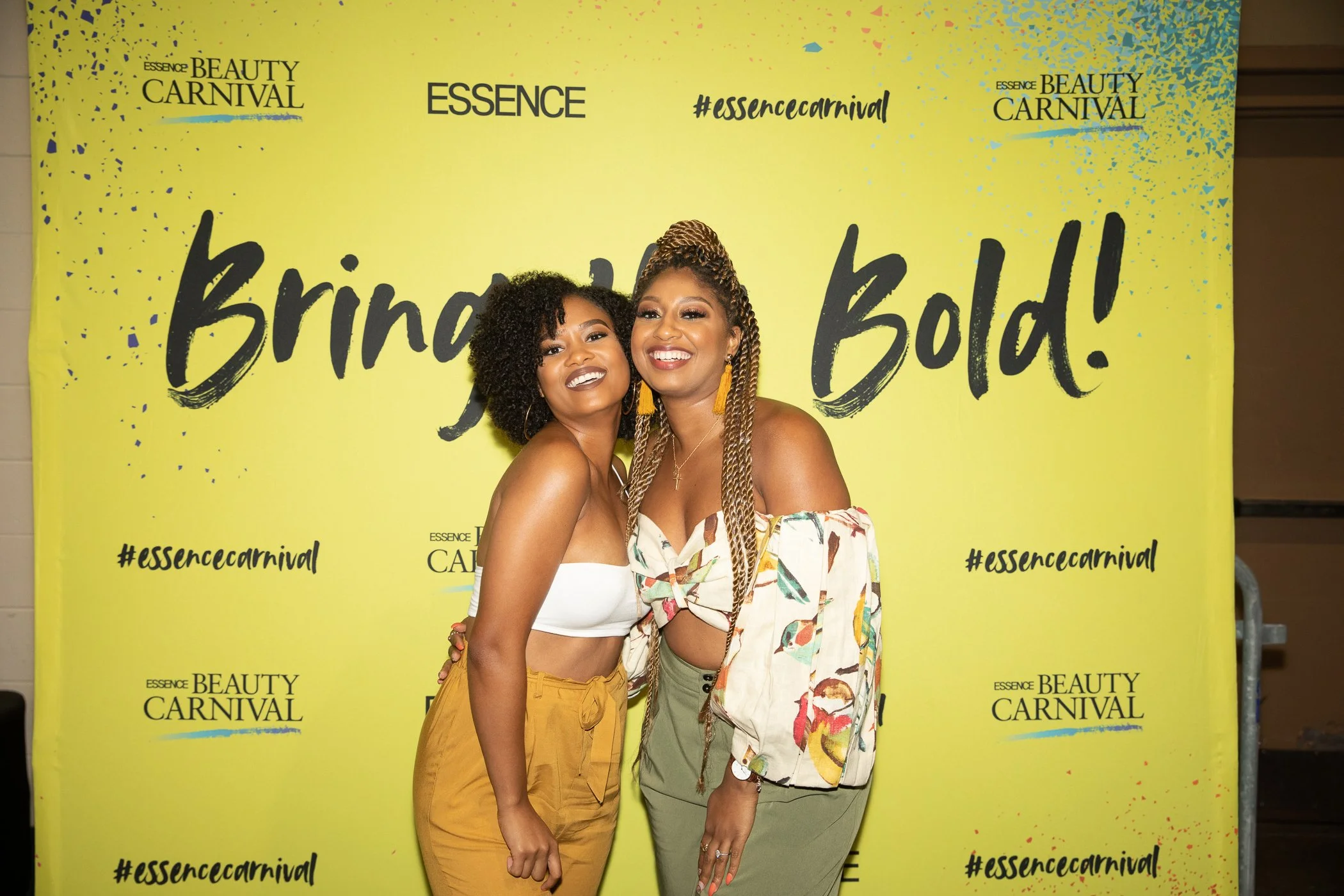 Two women smiling and posing in front of a yellow backdrop at the Essence Beauty Carnival event. One is wearing a white crop top and mustard yellow pants, the other is wearing a printed off-the-shoulder top and olive green pants.