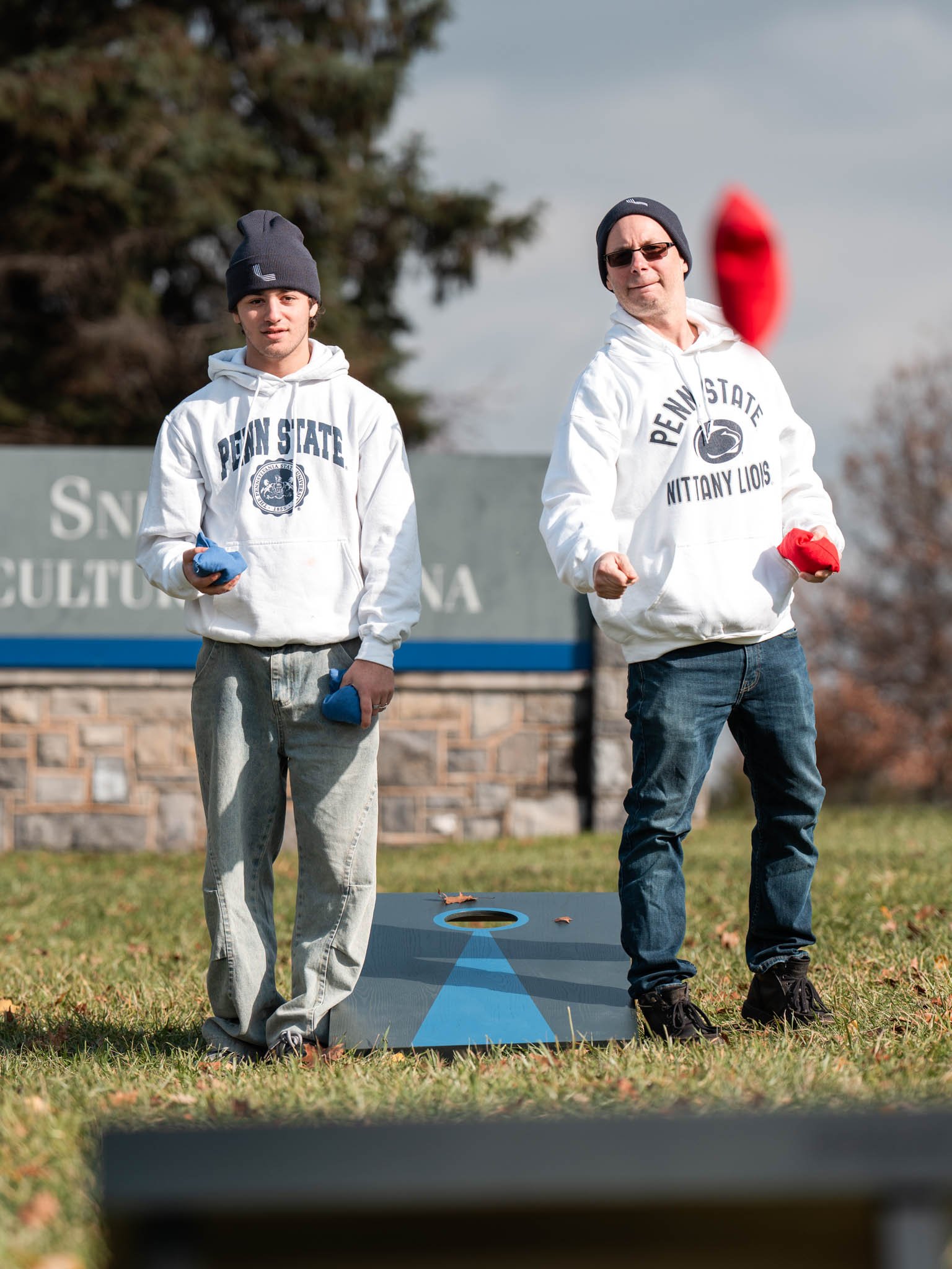 Two young men in Penn State hoodies and beanies playing cornhole on a grassy field, with trees and a sign in the background.