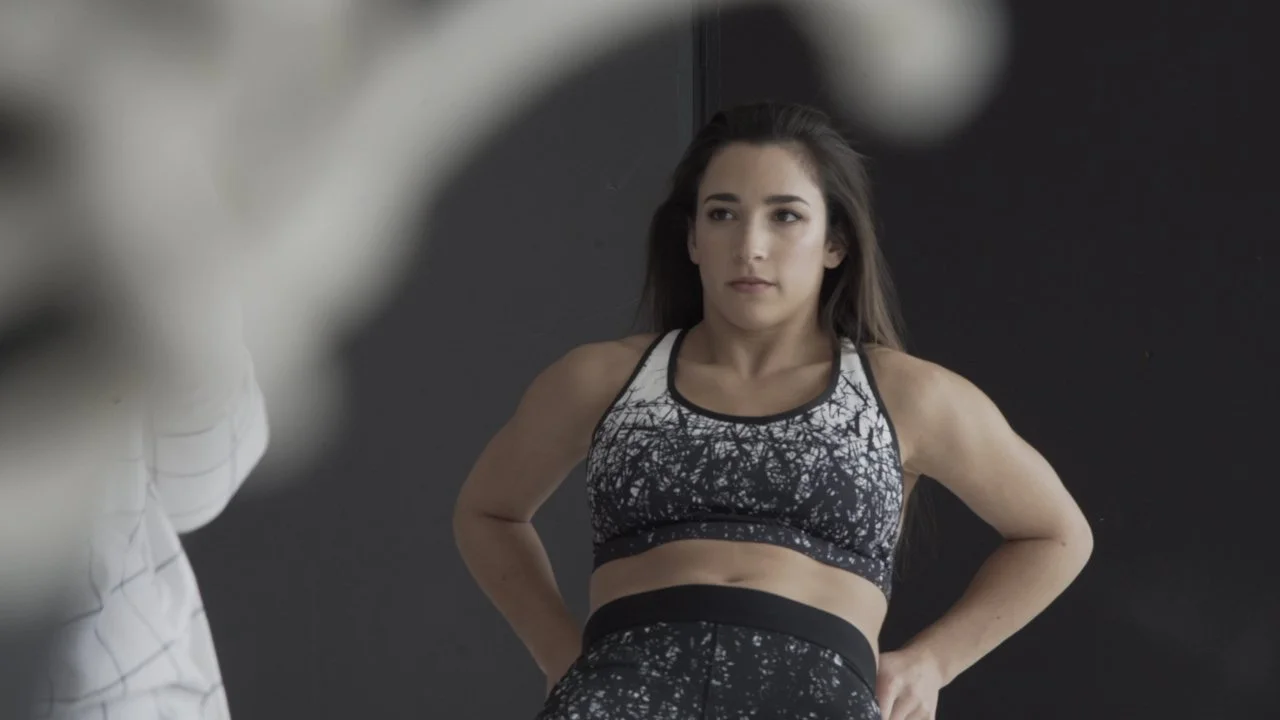 A woman with long brown hair in workout clothing stands with her hands on her hips, facing a person in the foreground who is out of focus.