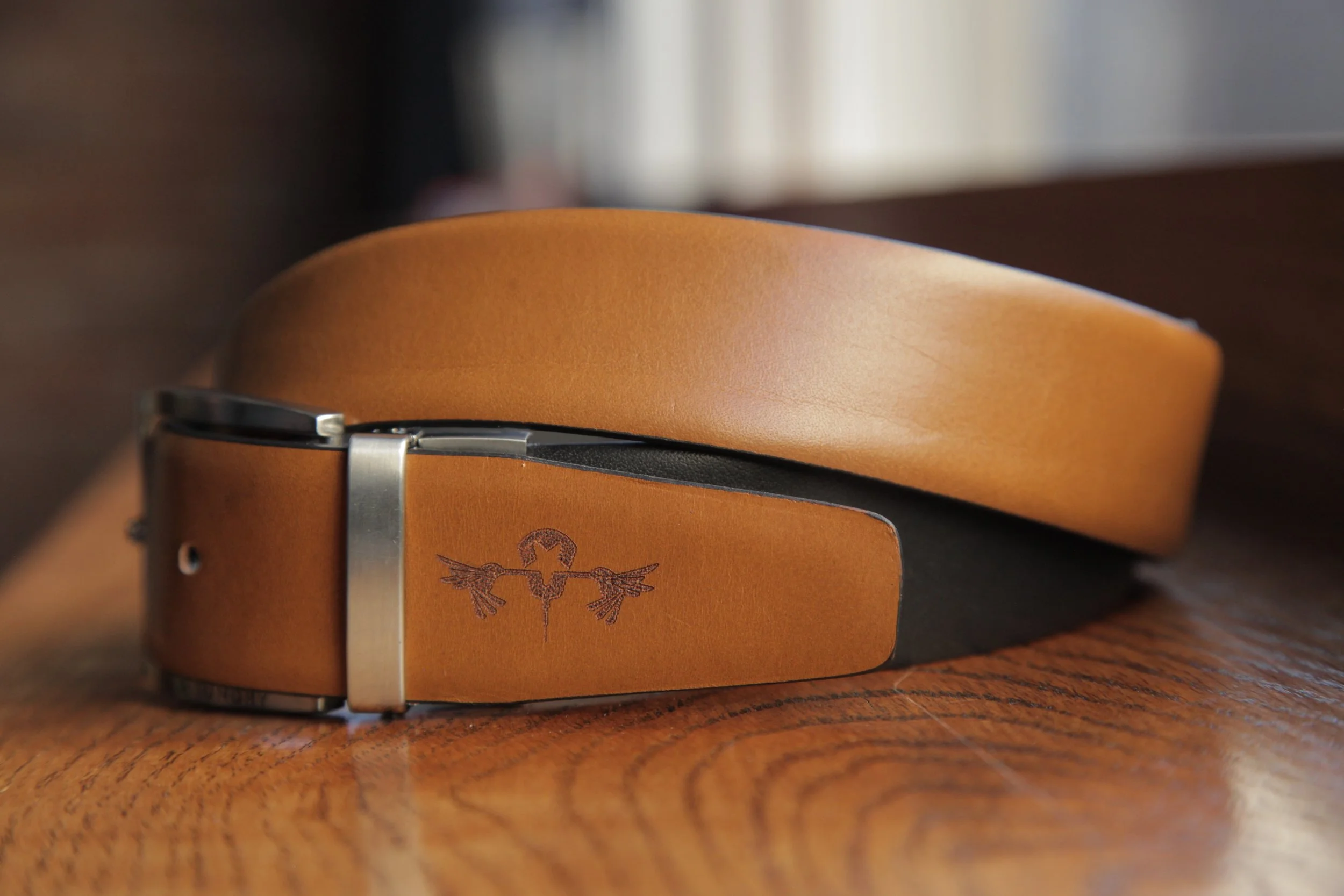 Leather belt with a metal buckle and engraved logo, resting on a wooden surface.