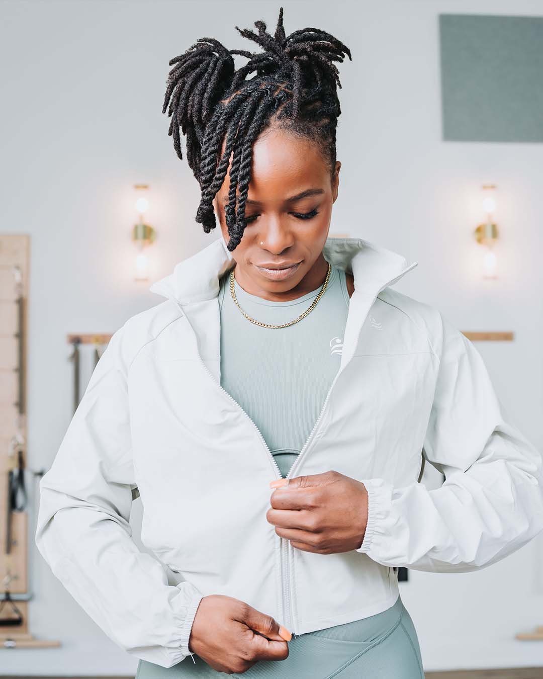 A woman with dreadlocks wearing a light gray sports outfit and a white zip-up jacket, standing in a room with minimalistic decor, looking down while adjusting her jacket.
