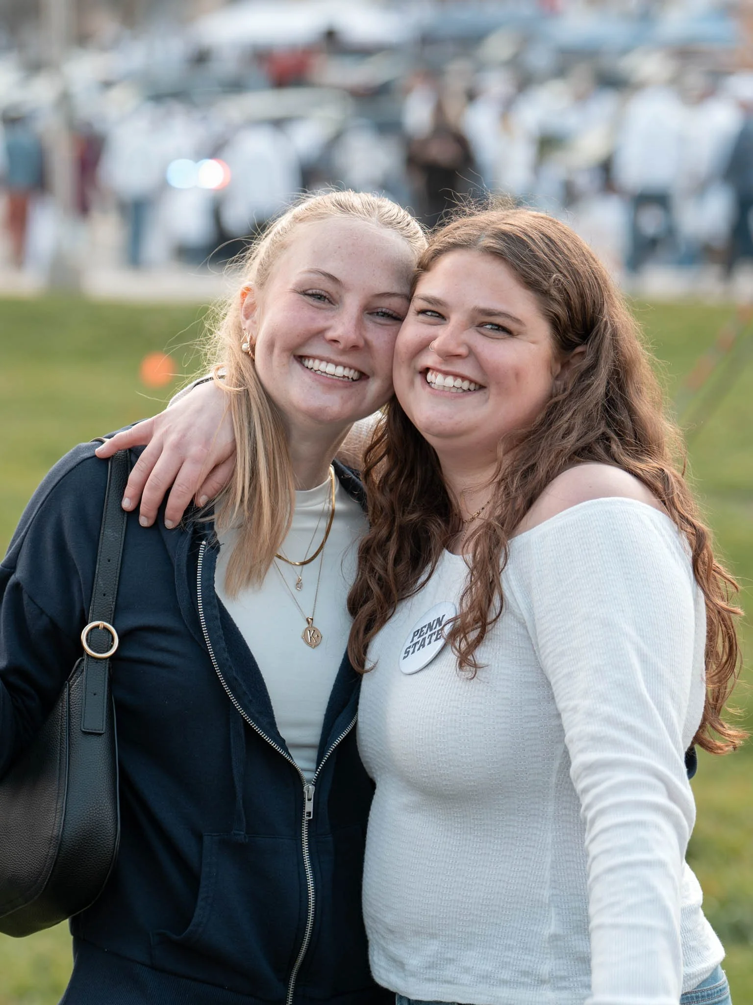 Two women smiling and embracing each other outdoors with a blurred background of a crowd and parking lot.