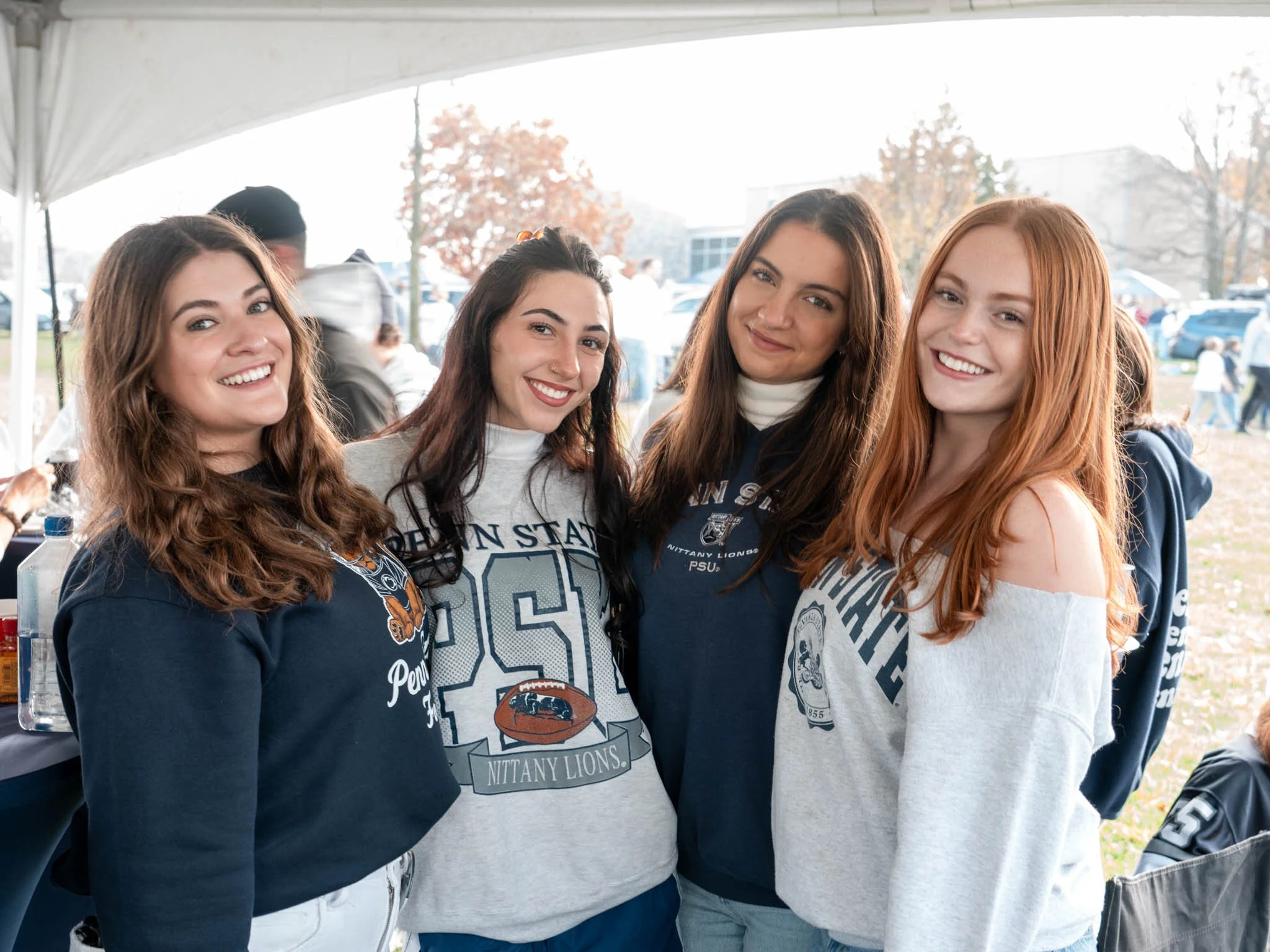 Four young women standing under a tent outdoors, smiling at the camera, wearing Penn State and Penn State Nittany Lions apparel.