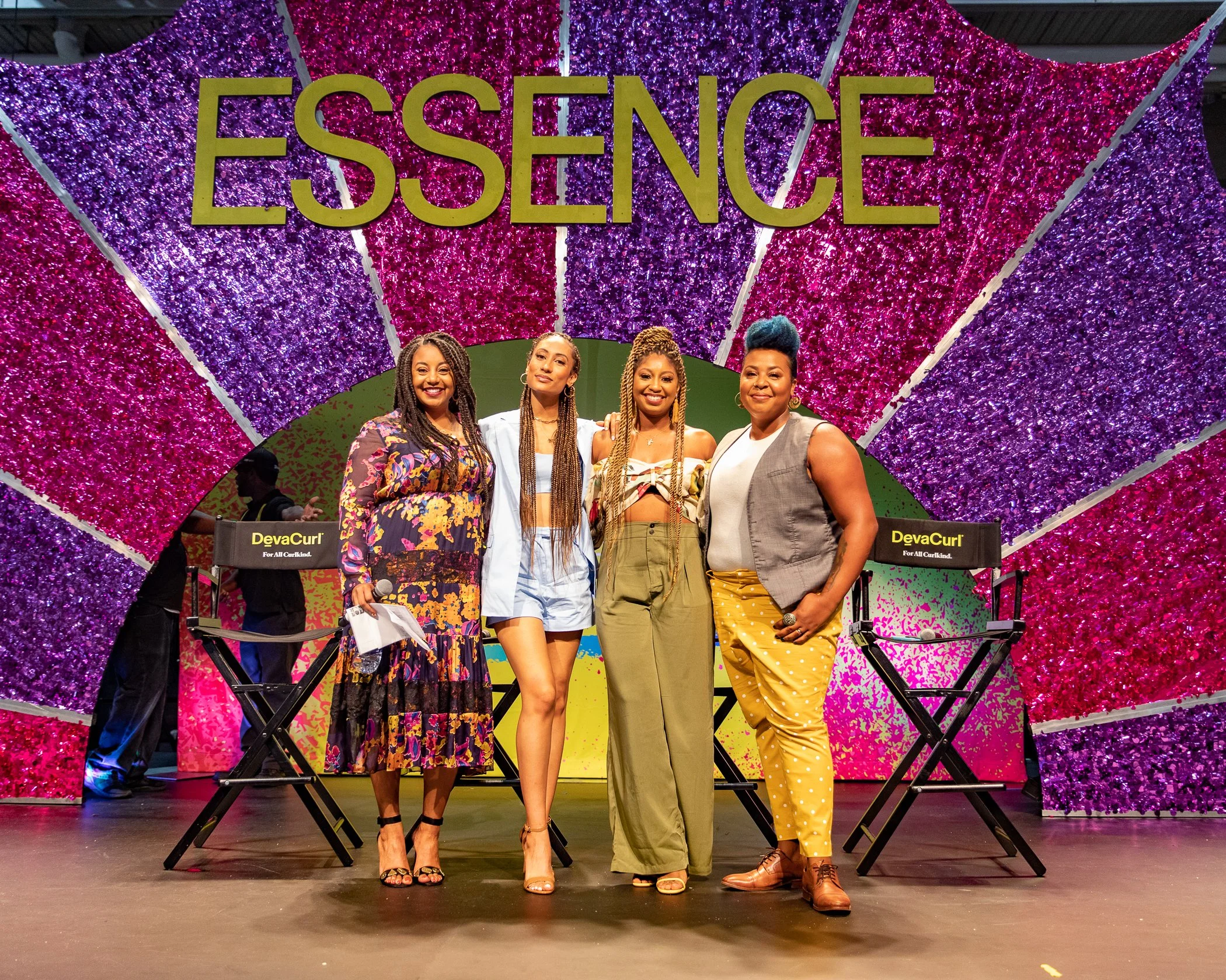 Four women standing on a stage with a colorful, purple and pink sparkling backdrop that spells out 'ESSENCE'. They are smiling and posing for a photo, with two director's chairs labeled 'DevaCurl' on either side of them.