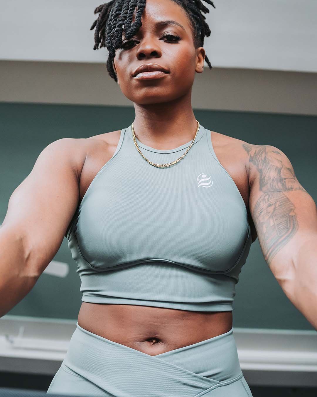 A woman wearing a light gray athletic sports bra and matching pants takes a selfie in a gym, showing her upper body and a tattoo on her left arm.
