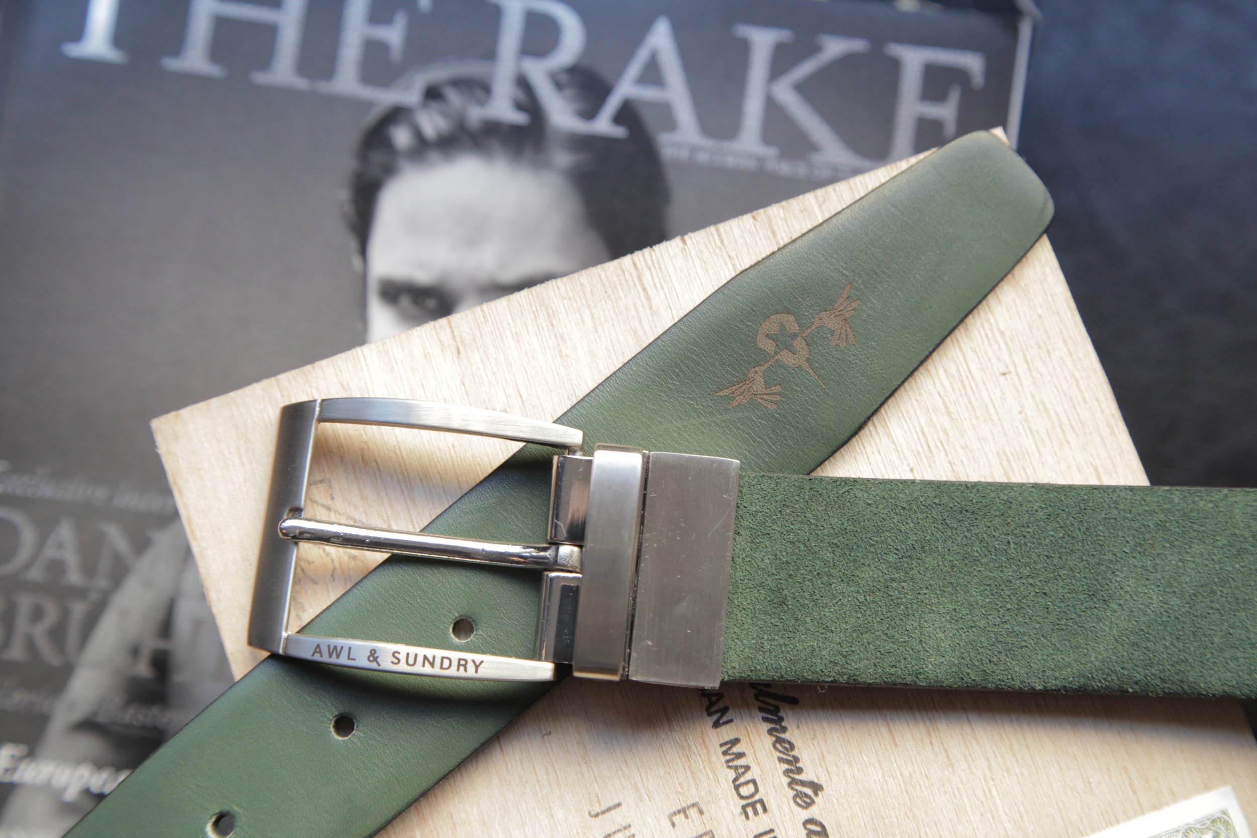 A green leather belt with a silver buckle, placed on top of a wooden surface with a magazine or book in the background. The belt has the words "AWL & SUNDRY" engraved on the buckle.