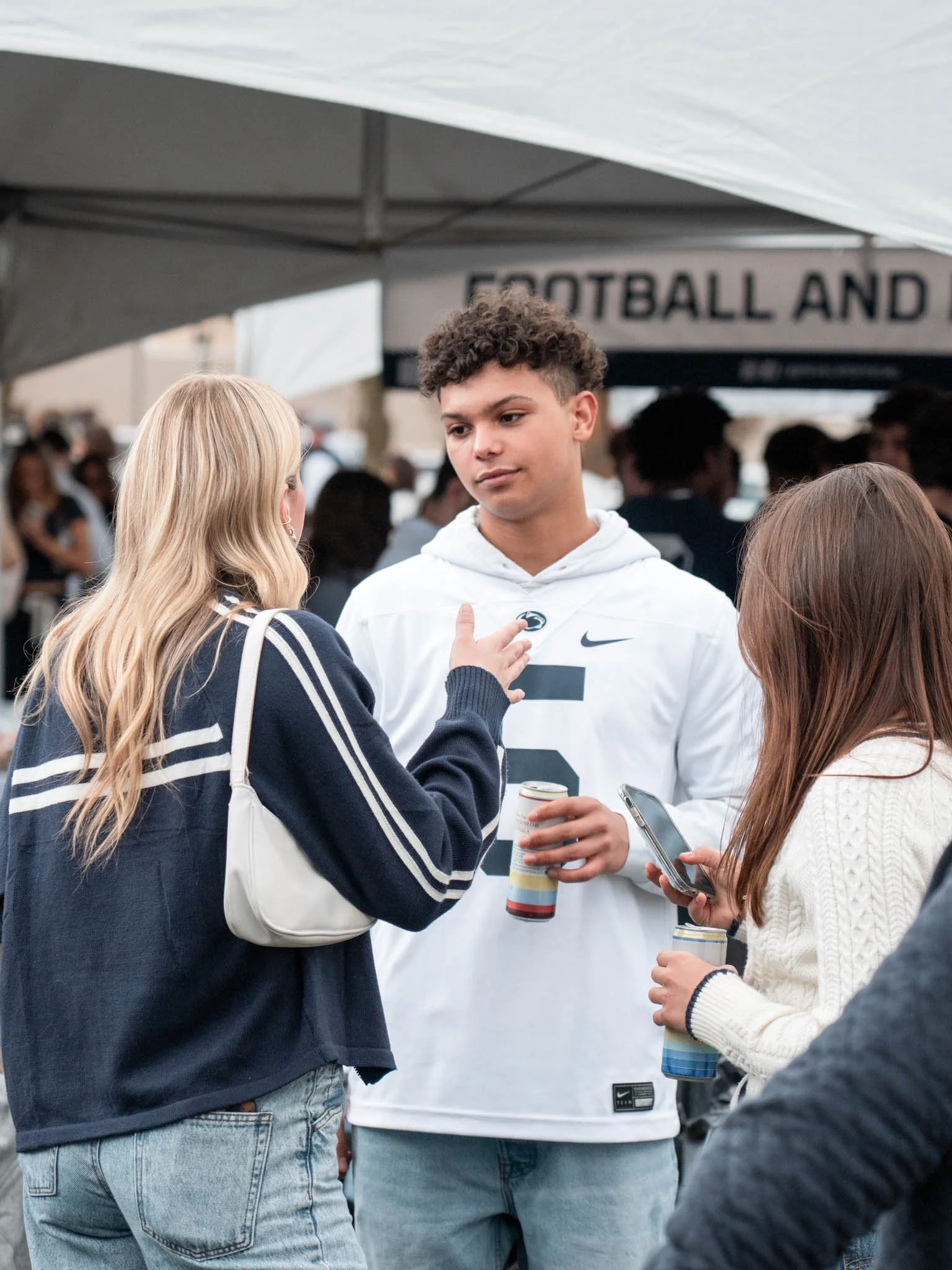 Three young people, two women and one man, are engaged in conversation at an outdoor event. The man is wearing a white sports jersey with the number 5 and is holding a drink. One woman, with blonde hair, is touching the man's chest and facing him. Th