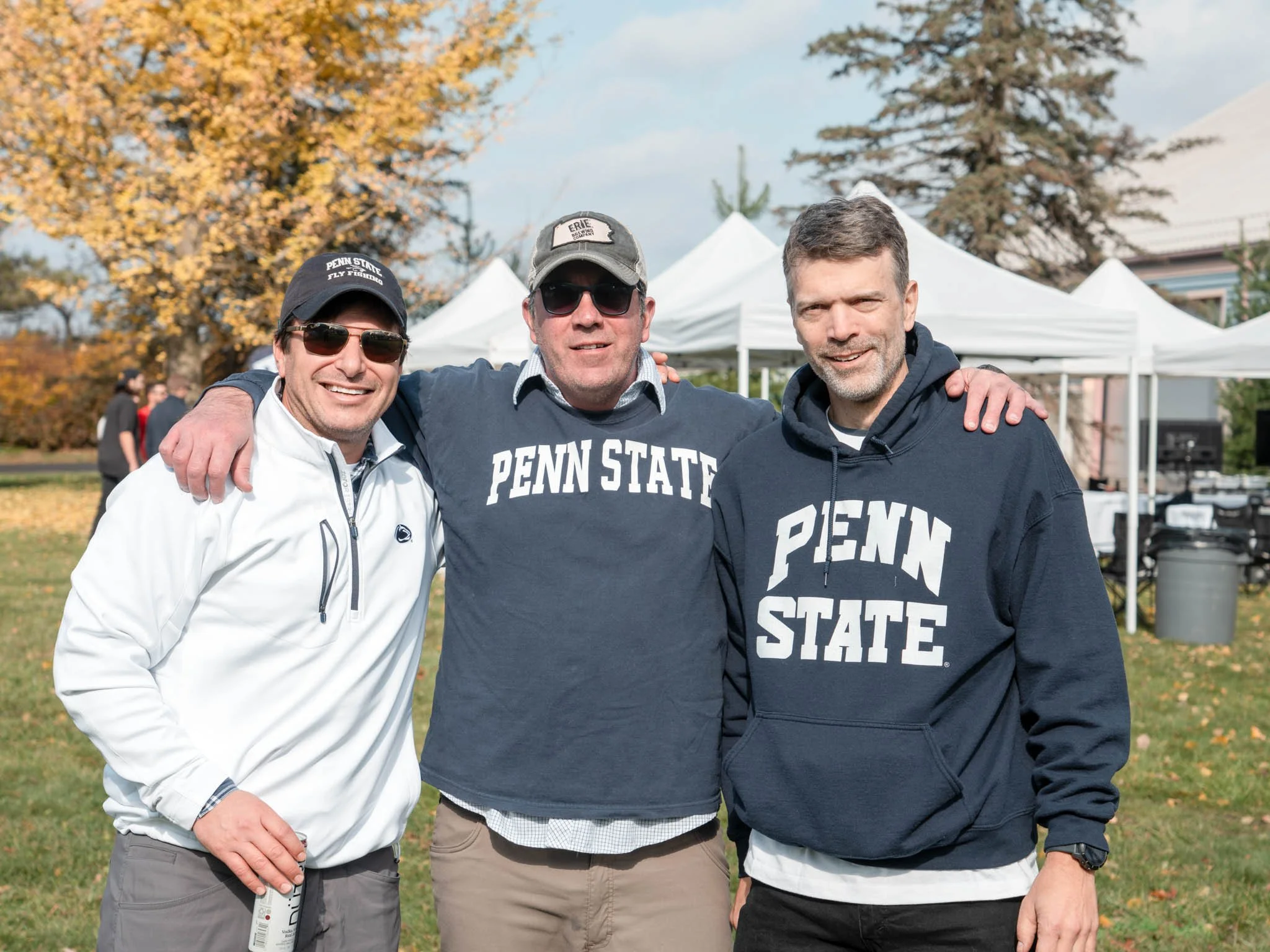 Three men wearing Penn State apparel standing outdoors at a fall event, smiling for the camera with tents and trees with colorful leaves in the background.