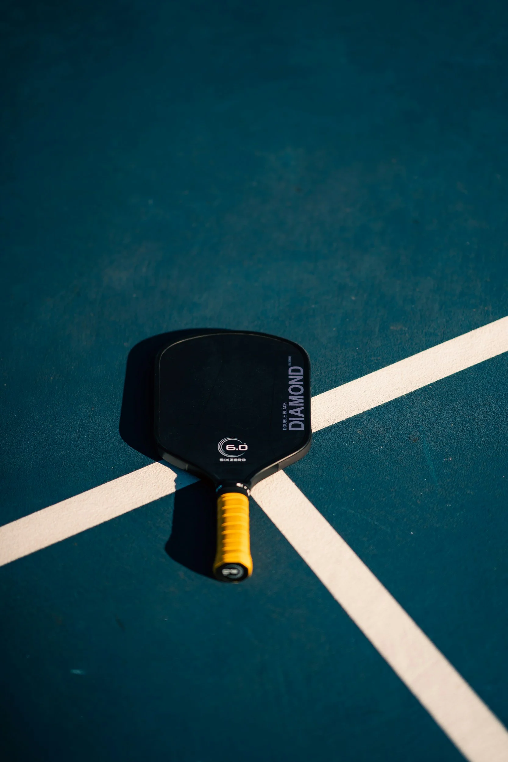 A black padel racket with yellow grip resting on a blue tennis court near white court lines.