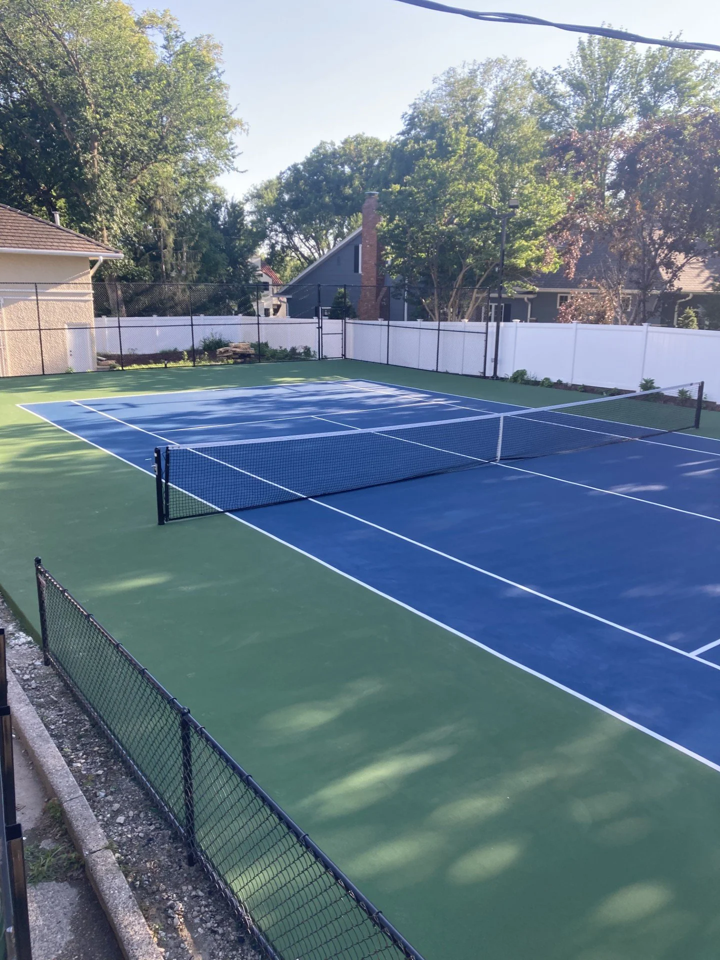 Outdoor tennis court with blue playing surface, surrounded by white fence, with trees and houses in background on a sunny day.