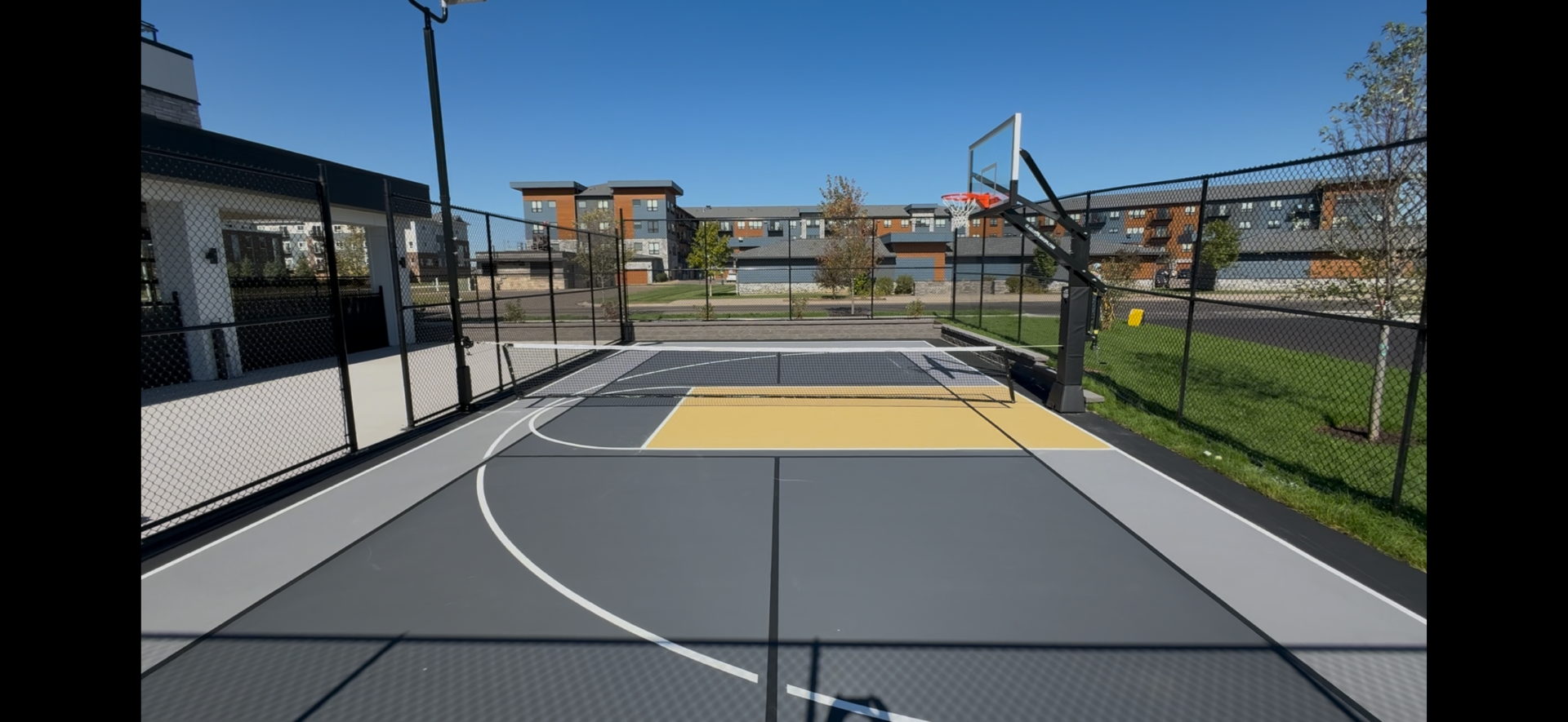 An outdoor basketball court with a basketball hoop and a fenced perimeter, set against a backdrop of residential buildings and a clear blue sky.