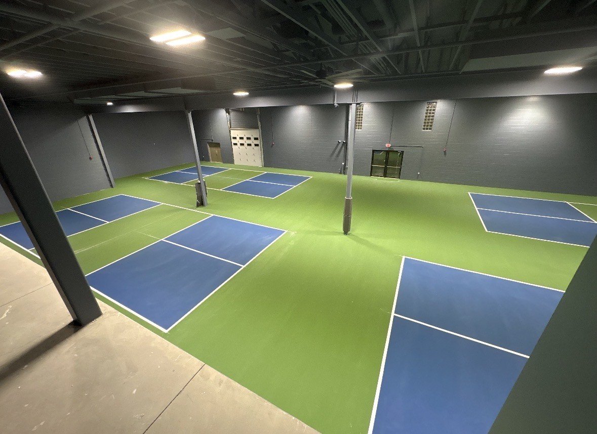 Indoor pickleball court with green flooring, blue court surfaces, and black walls. Several courts with white boundary lines are visible, and the ceiling has exposed black piping and lighting fixtures.