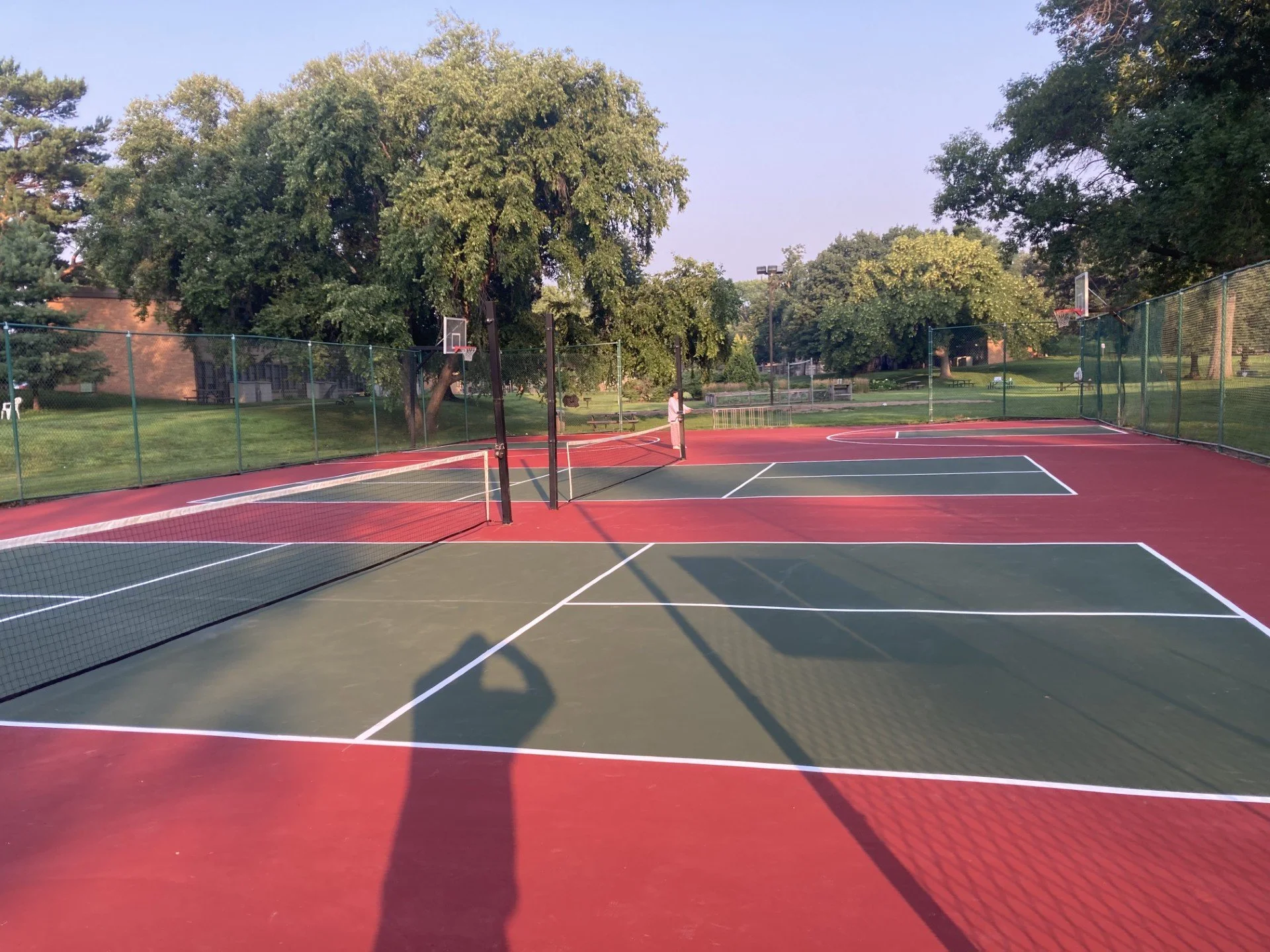 Empty outdoor tennis courts with a basketball court in the background, surrounded by trees and a grassy park area, during daytime.