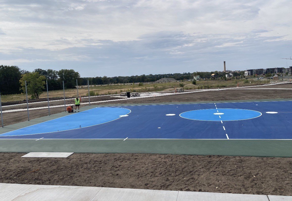 Newly painted outdoor basketball court in blue with white lines, surrounded by dirt and with a person working on it.