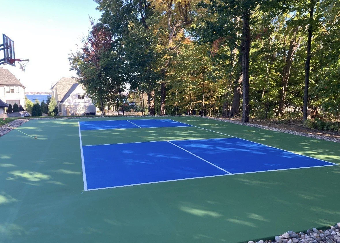 An outdoor tennis court surrounded by trees with houses in the background, a basketball hoop on the left, and the court surface is green with blue playing areas.