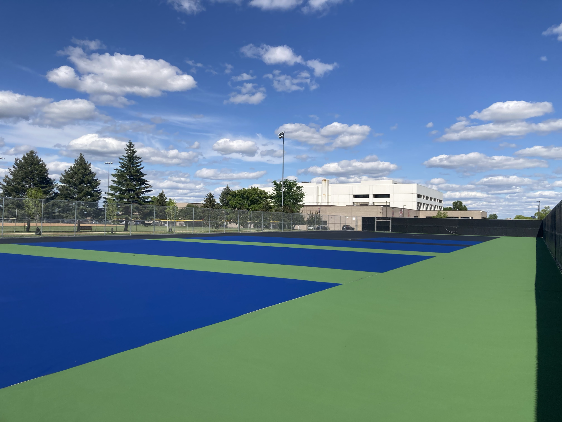 An outdoor tennis or pickleball court with bright blue and green surface colors, enclosed by a chain-link fence, with trees and a white building in the background under a partly cloudy sky.