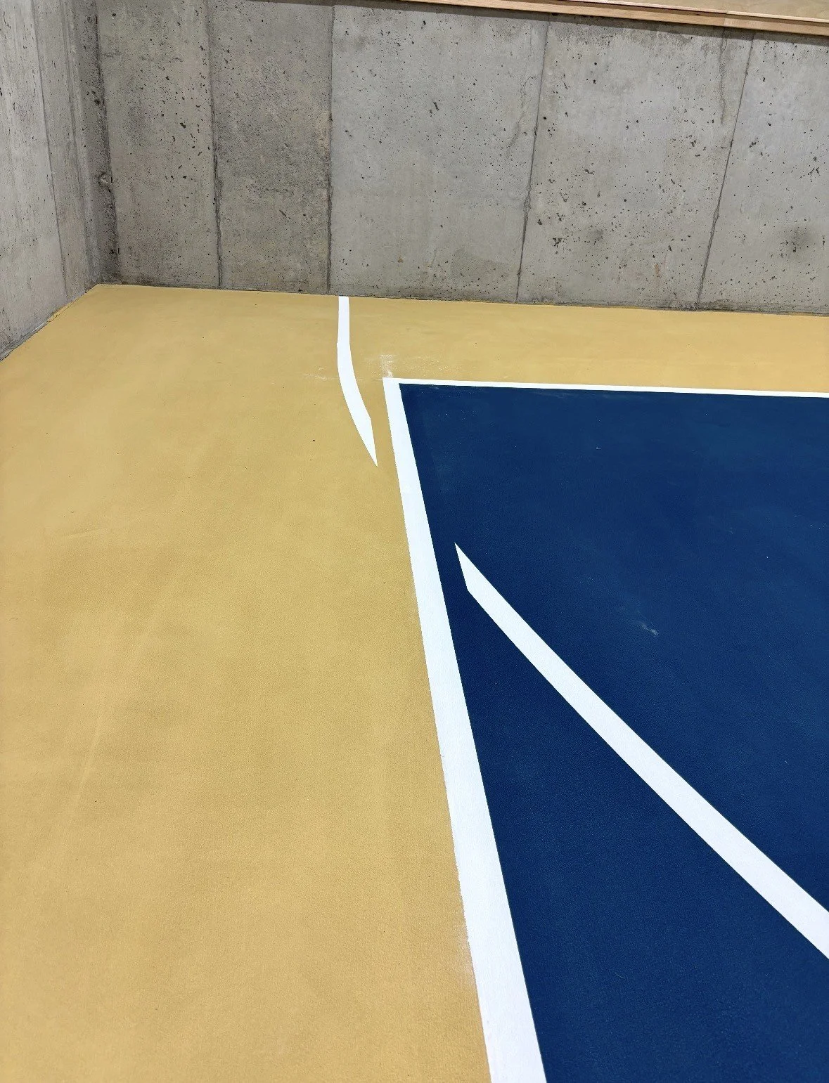 Close-up of a painted basketball court corner with yellow and blue sections separated by white lines, with concrete wall in the background.