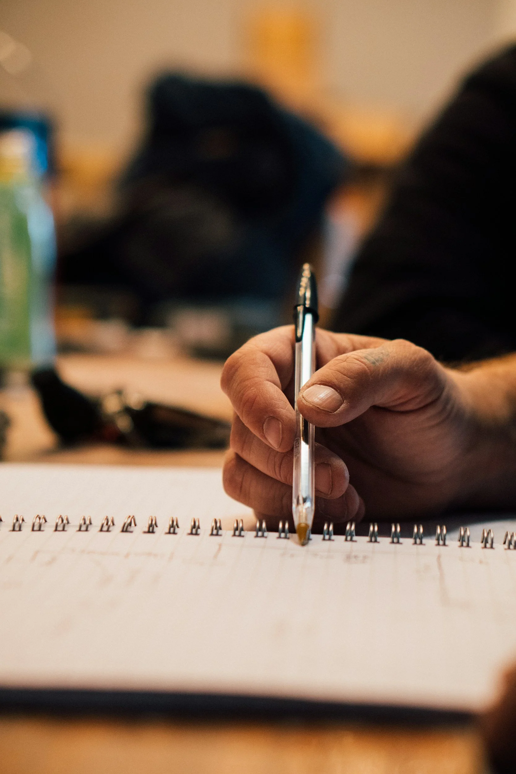 A person's hand holding a silver pen and writing on a spiral notebook with printed text.